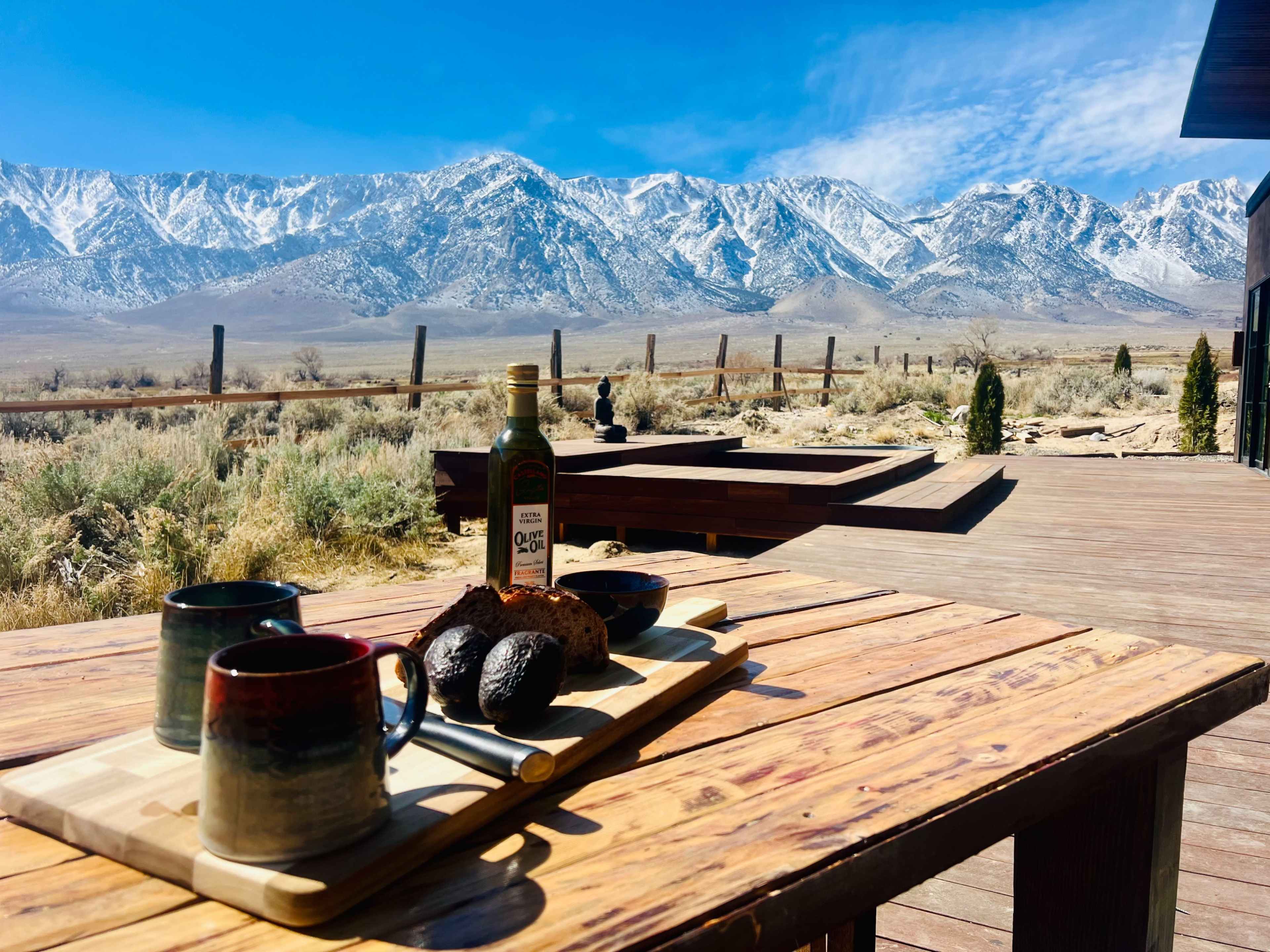A wooden table displays two mugs, a bottle of olive oil, and two avocados, with snow-capped mountains in the background under a clear blue sky.