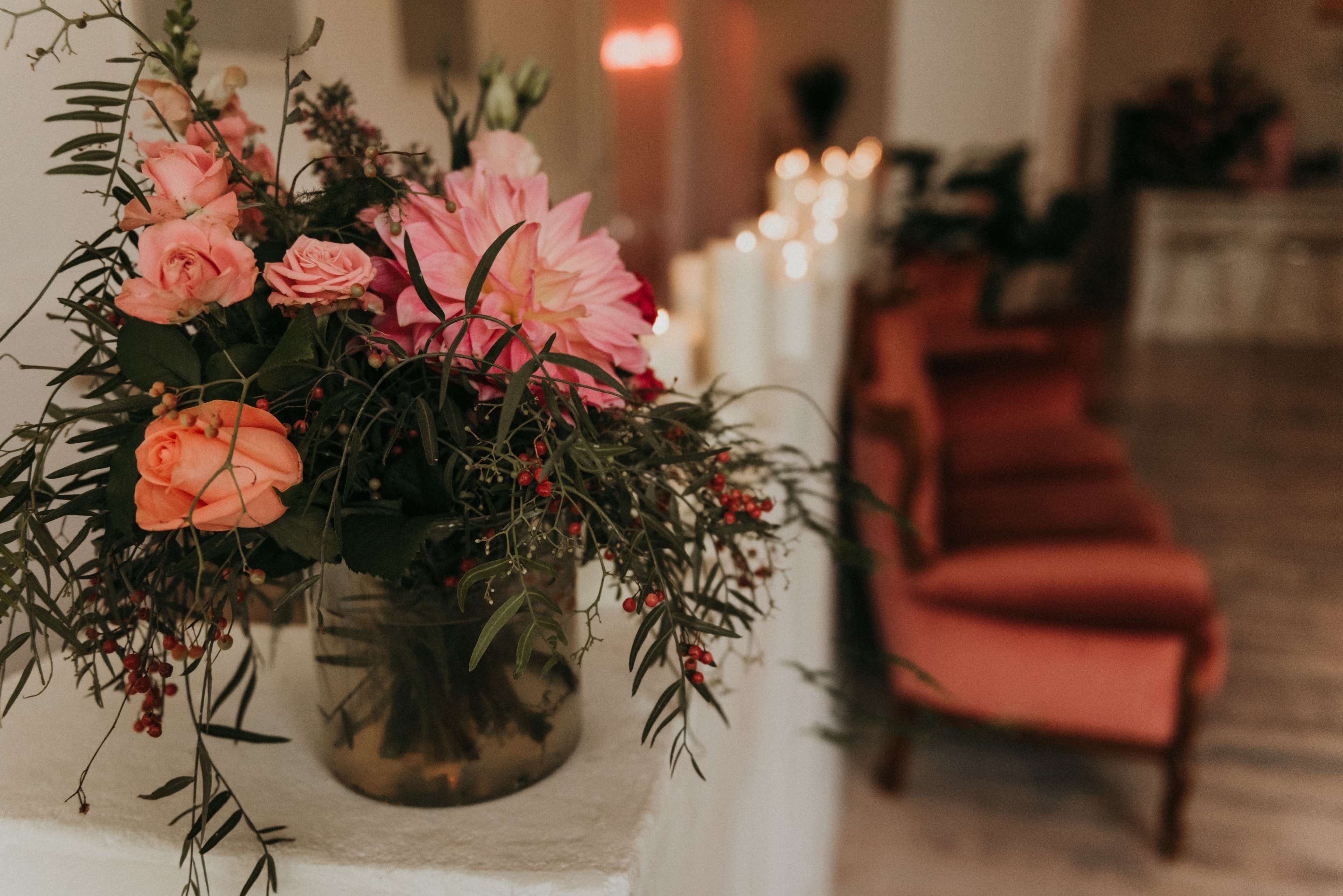 A vase of flowers sits on a ledge beside a row of pink armchairs and lit candles in a softly lit room.