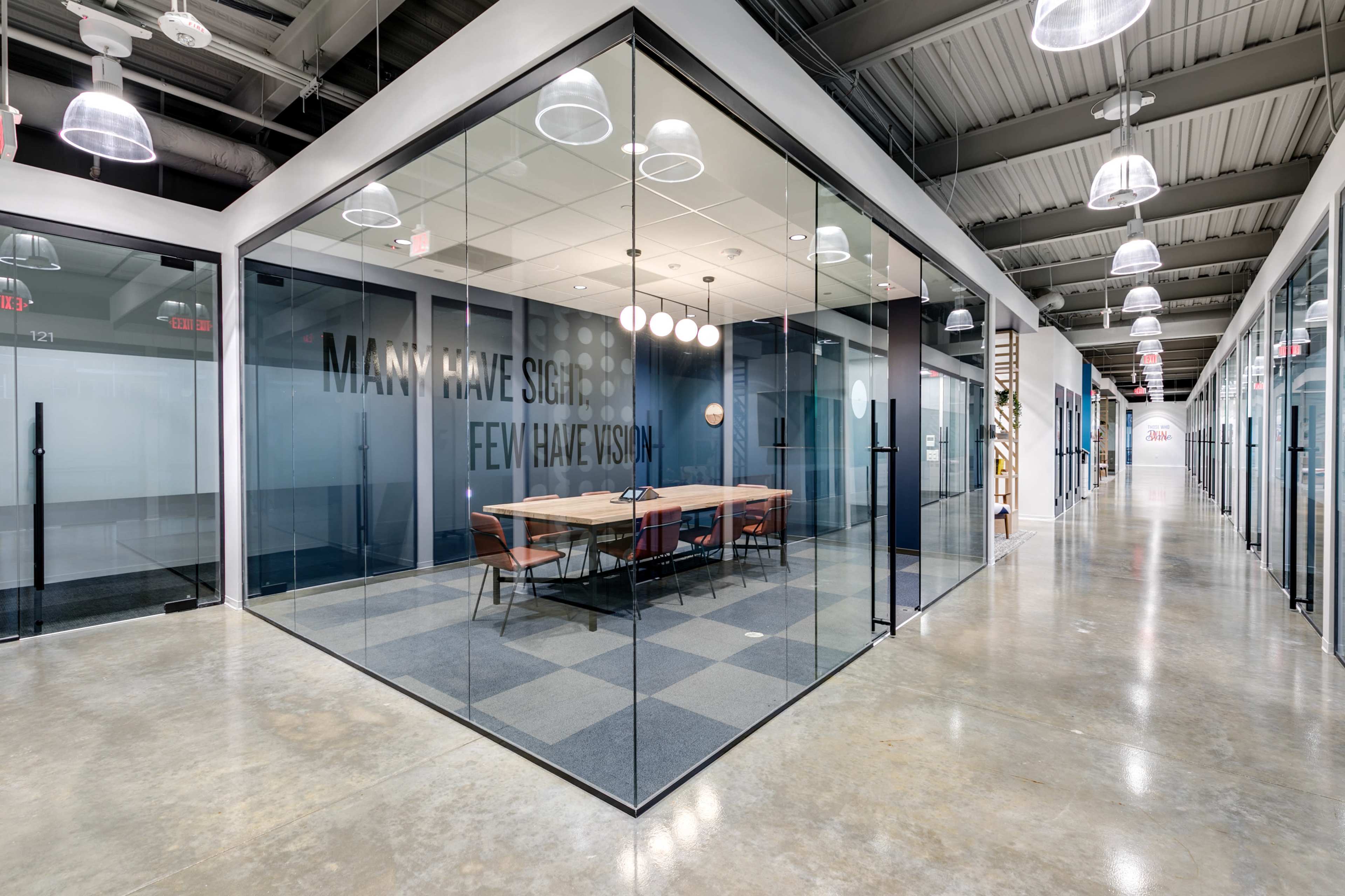 The image shows a modern office space featuring glass-walled conference rooms with a long table and a geometric floor pattern.