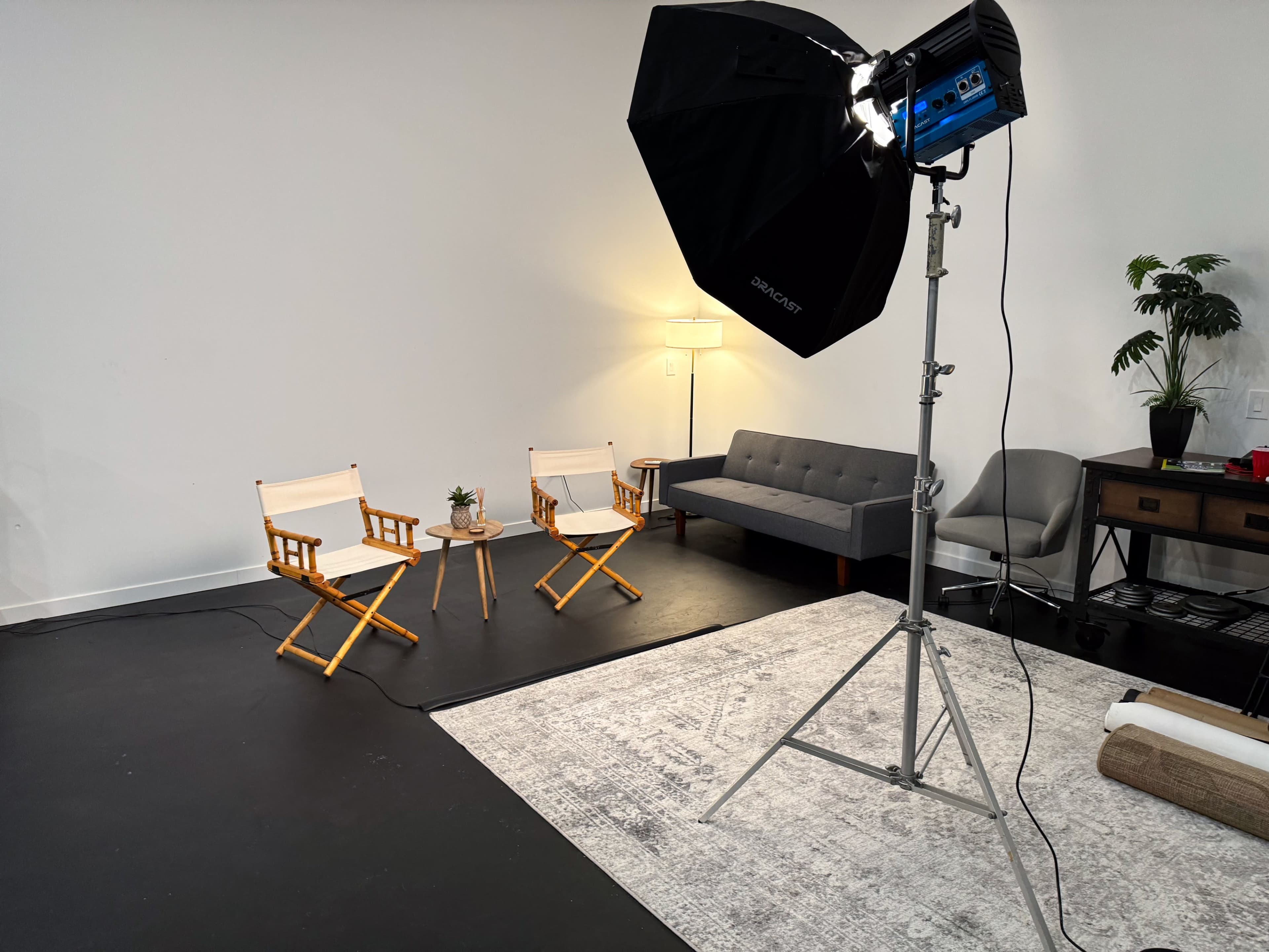 The image shows a well-lit studio space with two wooden chairs, a small table between them, and a couch in the background.