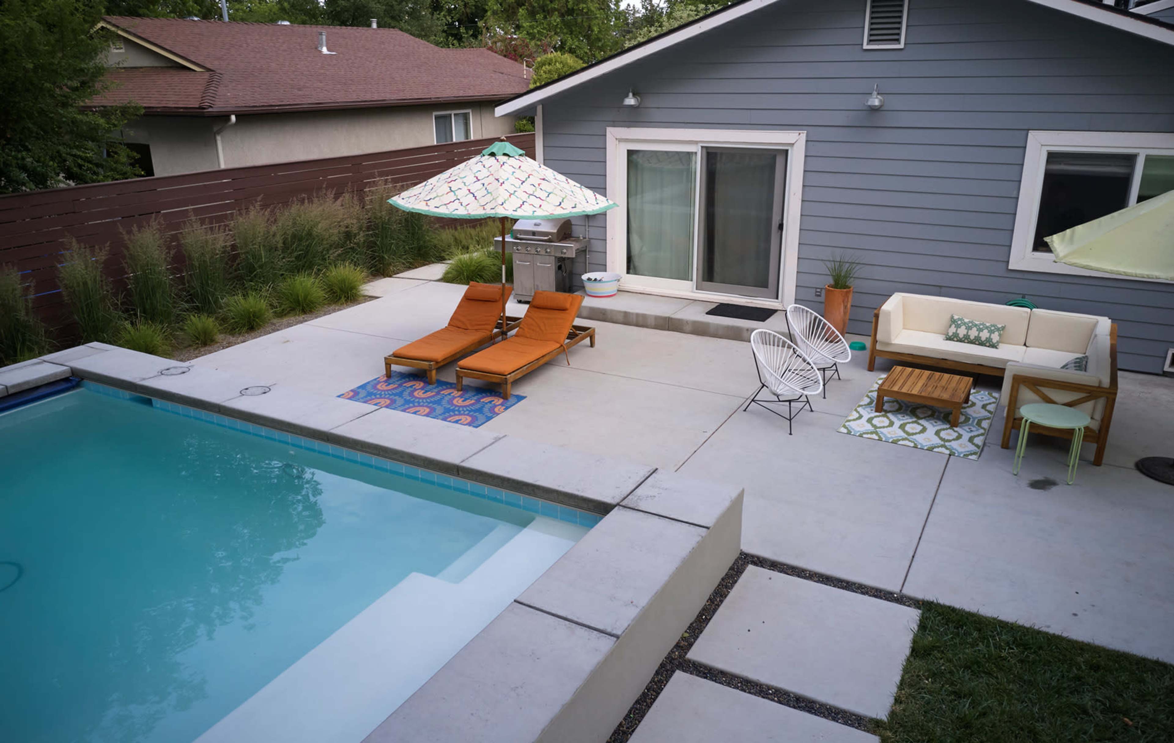 The image shows a backyard with a swimming pool, two orange lounge chairs with an umbrella, a grill, and patio furniture beside a gray house.