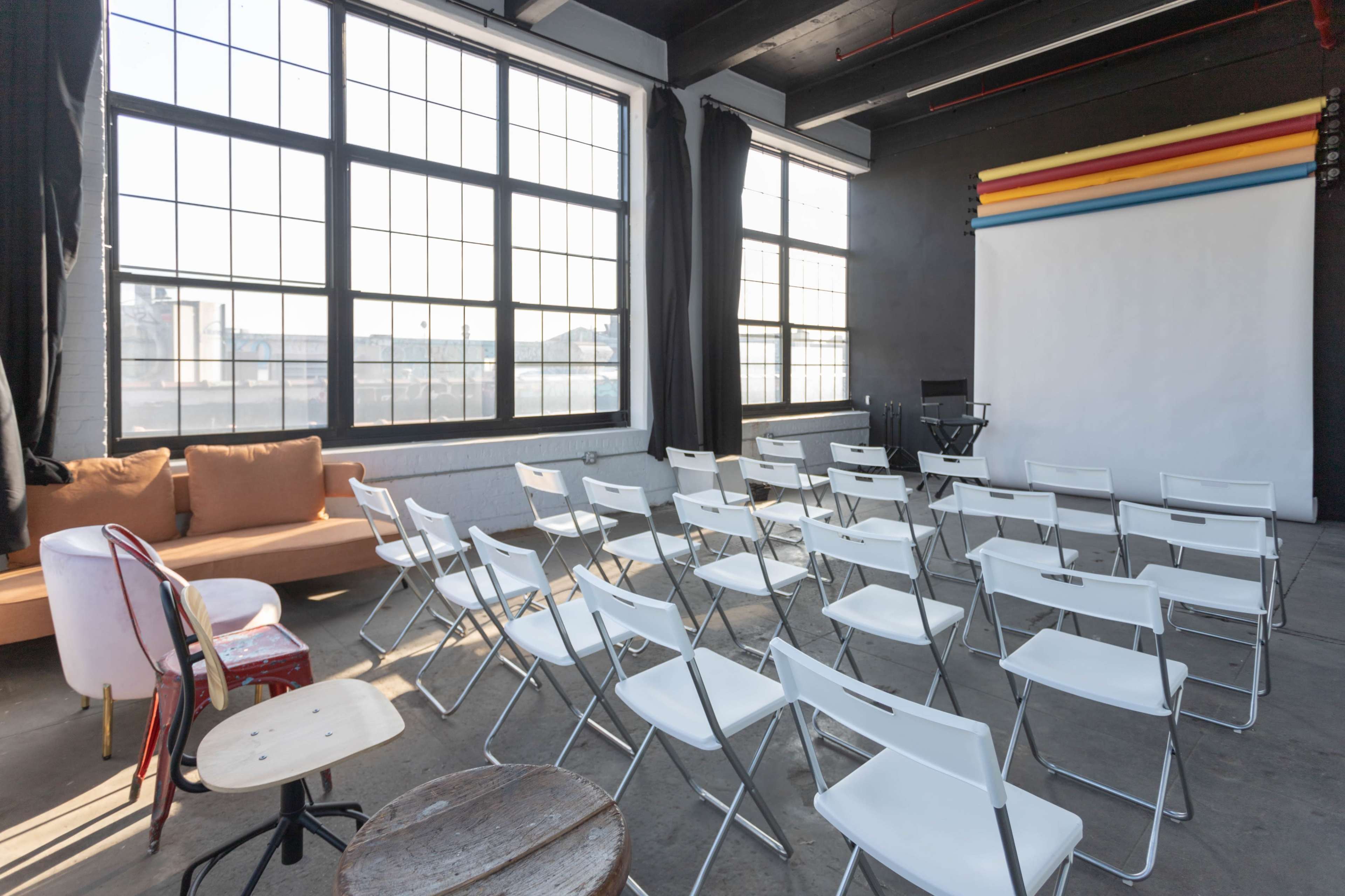 A spacious room with large windows contains rows of white folding chairs facing a blank backdrop, with a sofa and a wooden stool positioned nearby.