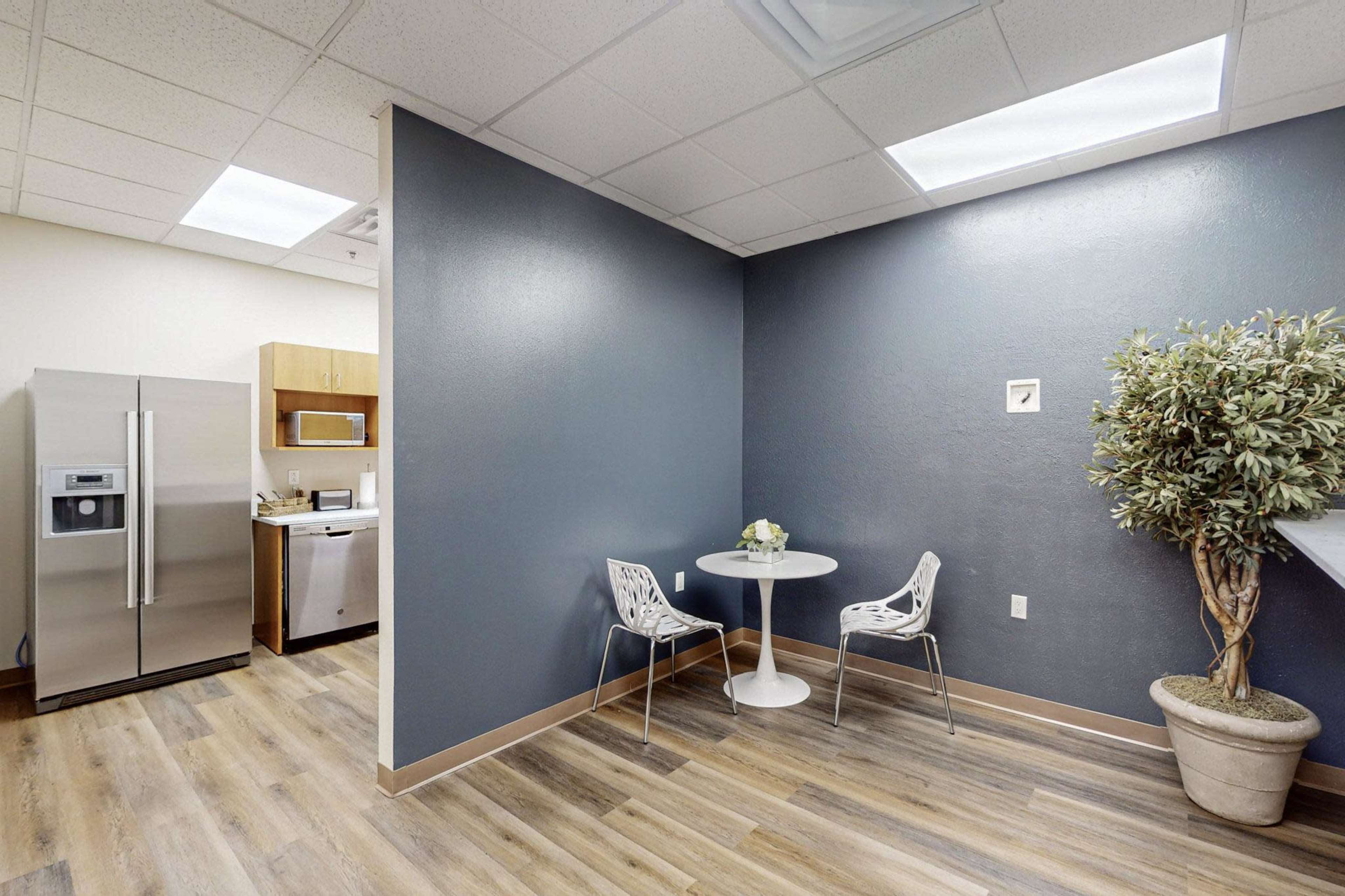 A modern kitchen area features a small round table with two chairs, a refrigerator, and a potted plant in the corner.