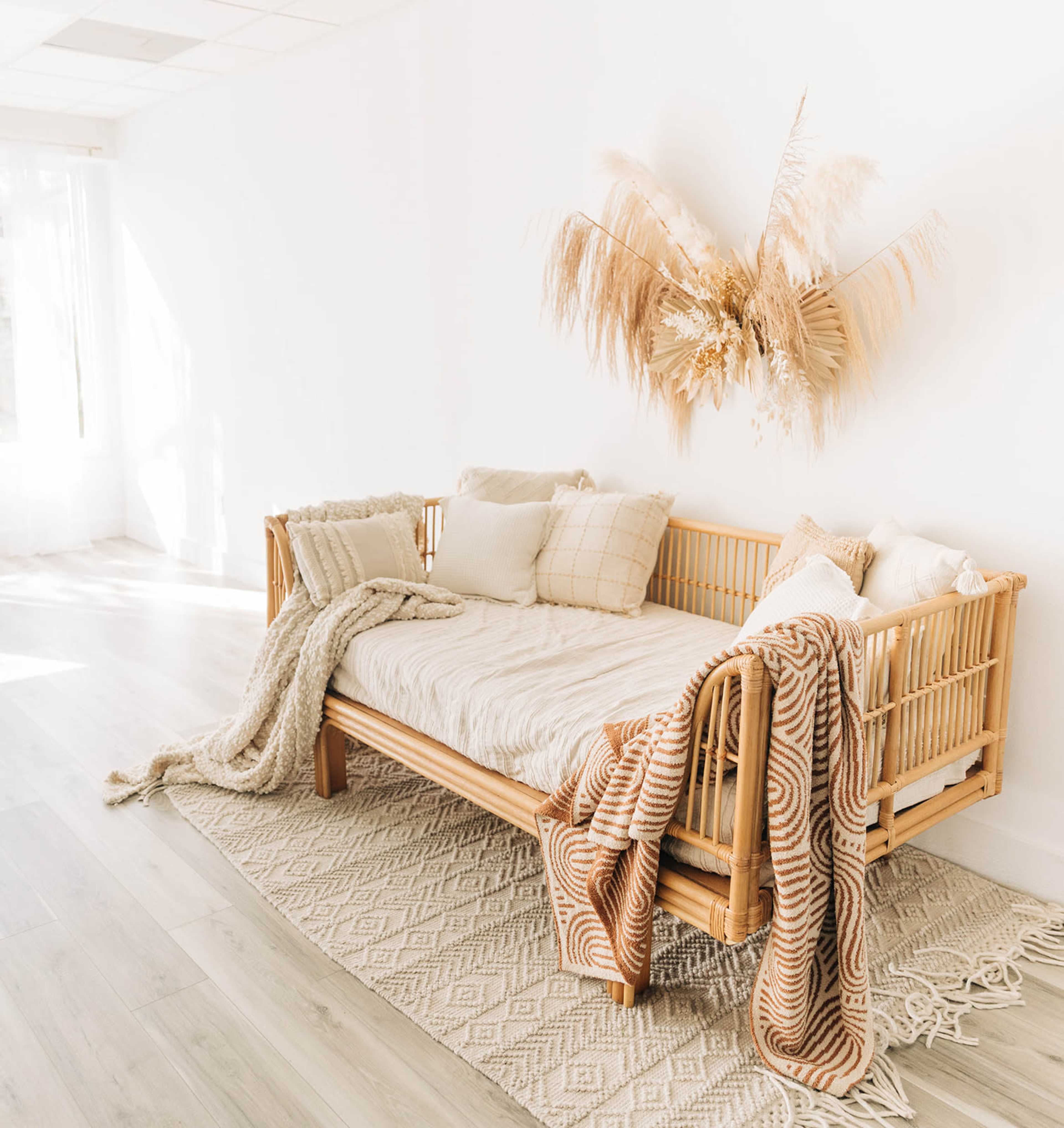 A rattan daybed is adorned with various cushions and a patterned throw, while decorative dried plants hang on the wall above it.