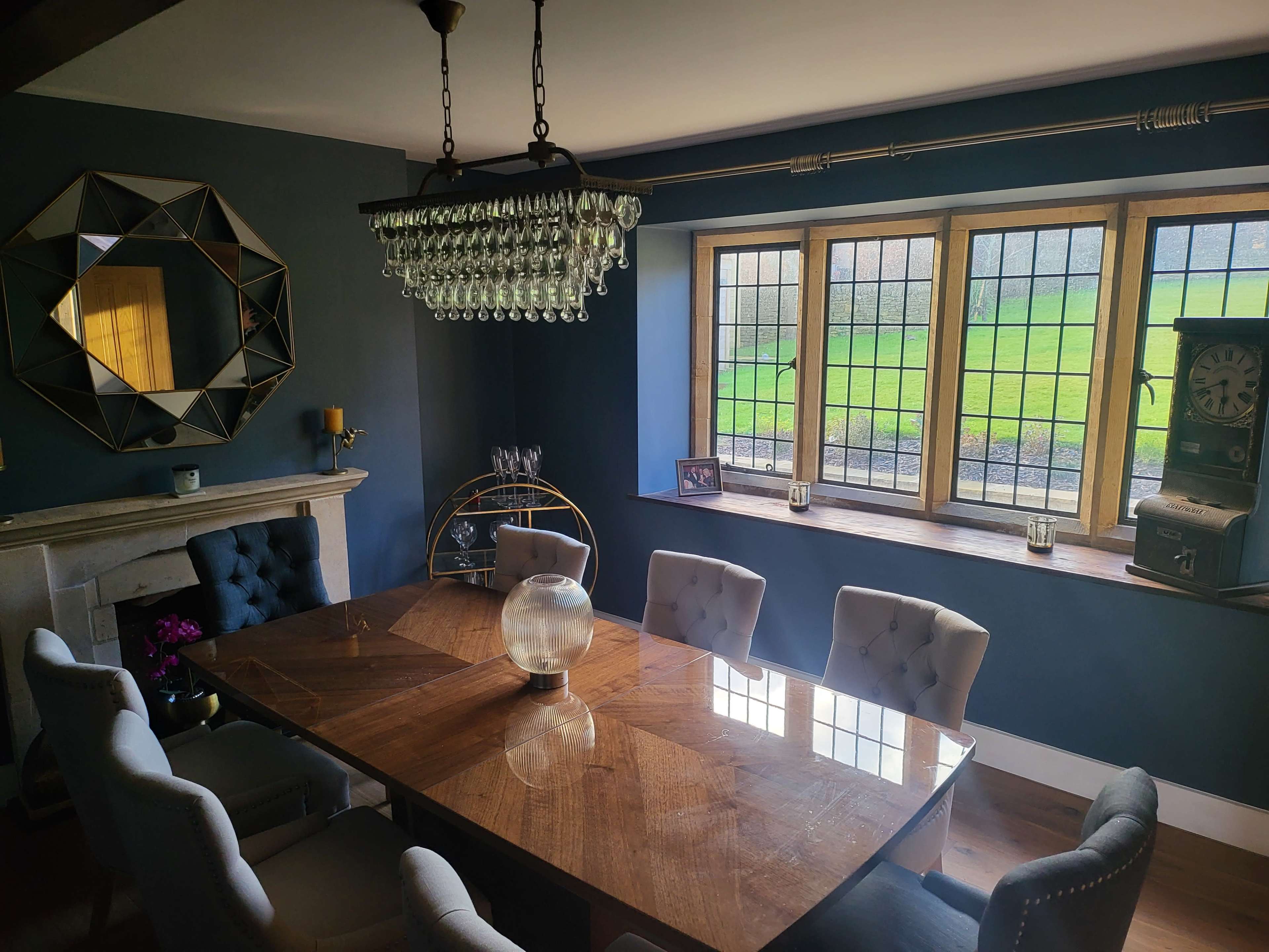A dining room with a wooden table, six upholstered chairs, a chandelier, and large windows overlooking a green landscape.