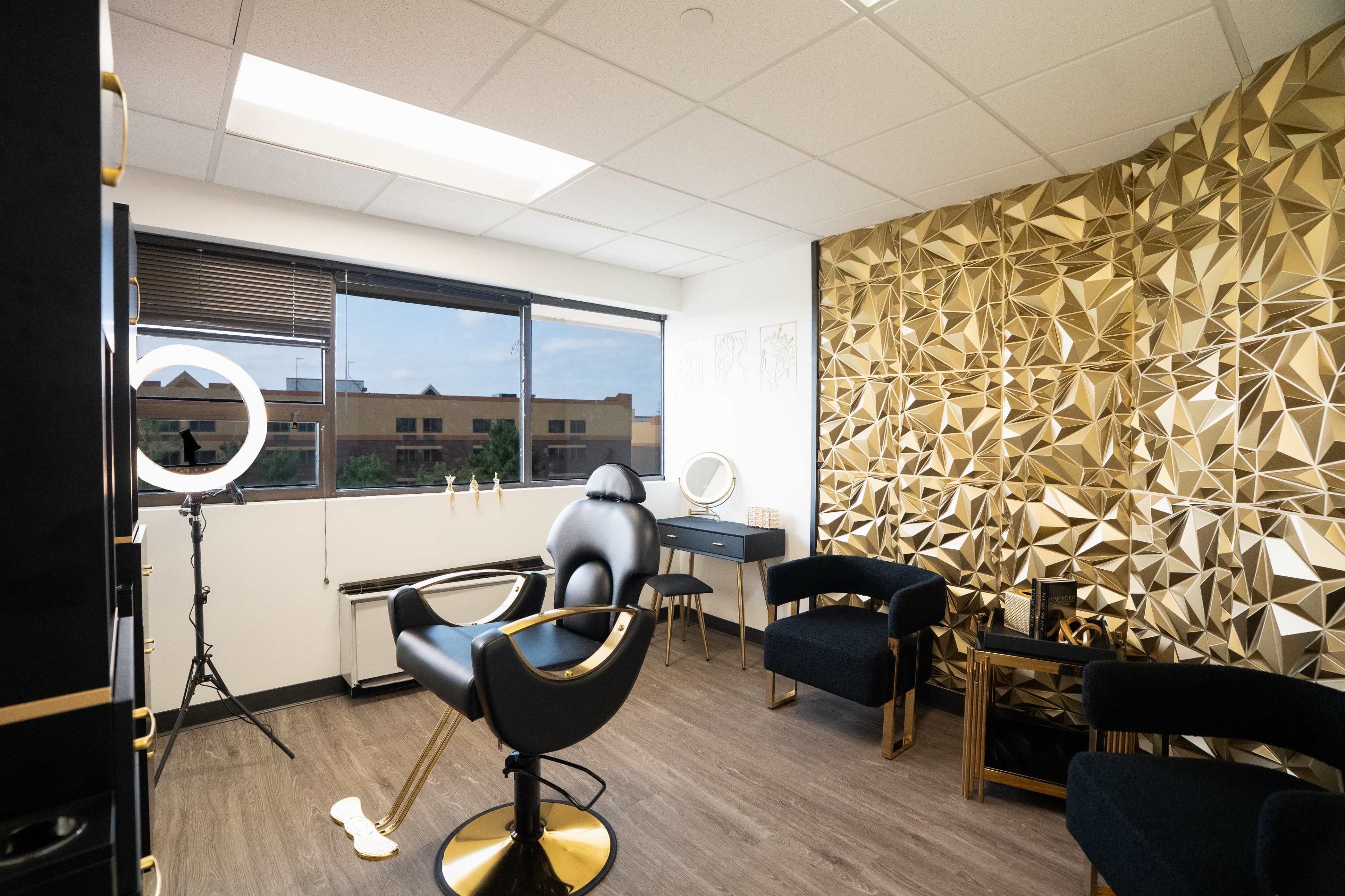 The image shows a modern beauty studio featuring a black swivel chair, a ring light, and a decorative golden wall.