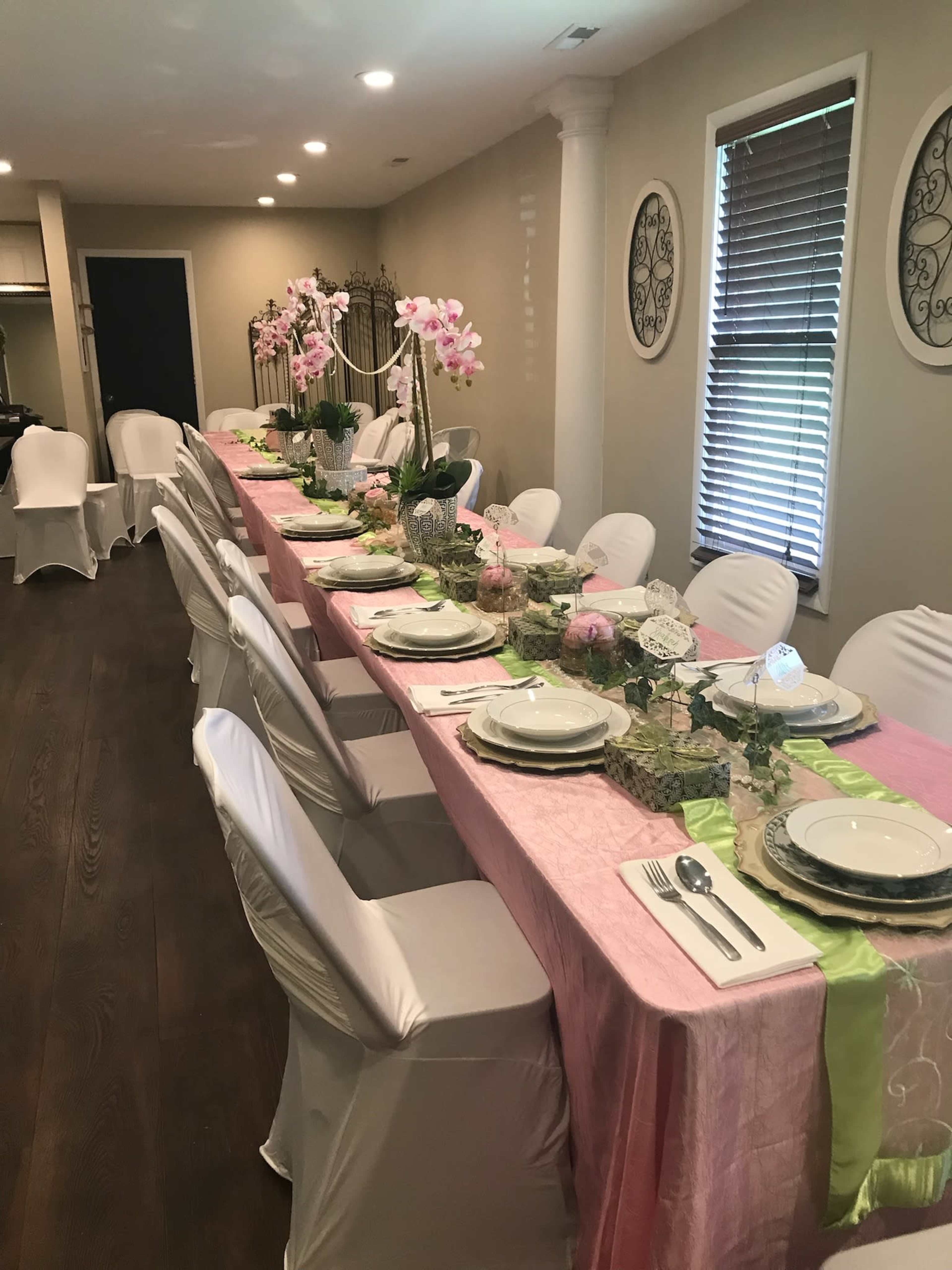 A long dining table is set with pink and green tablecloths, white chairs, and floral decorations, arranged for an elegant gathering.