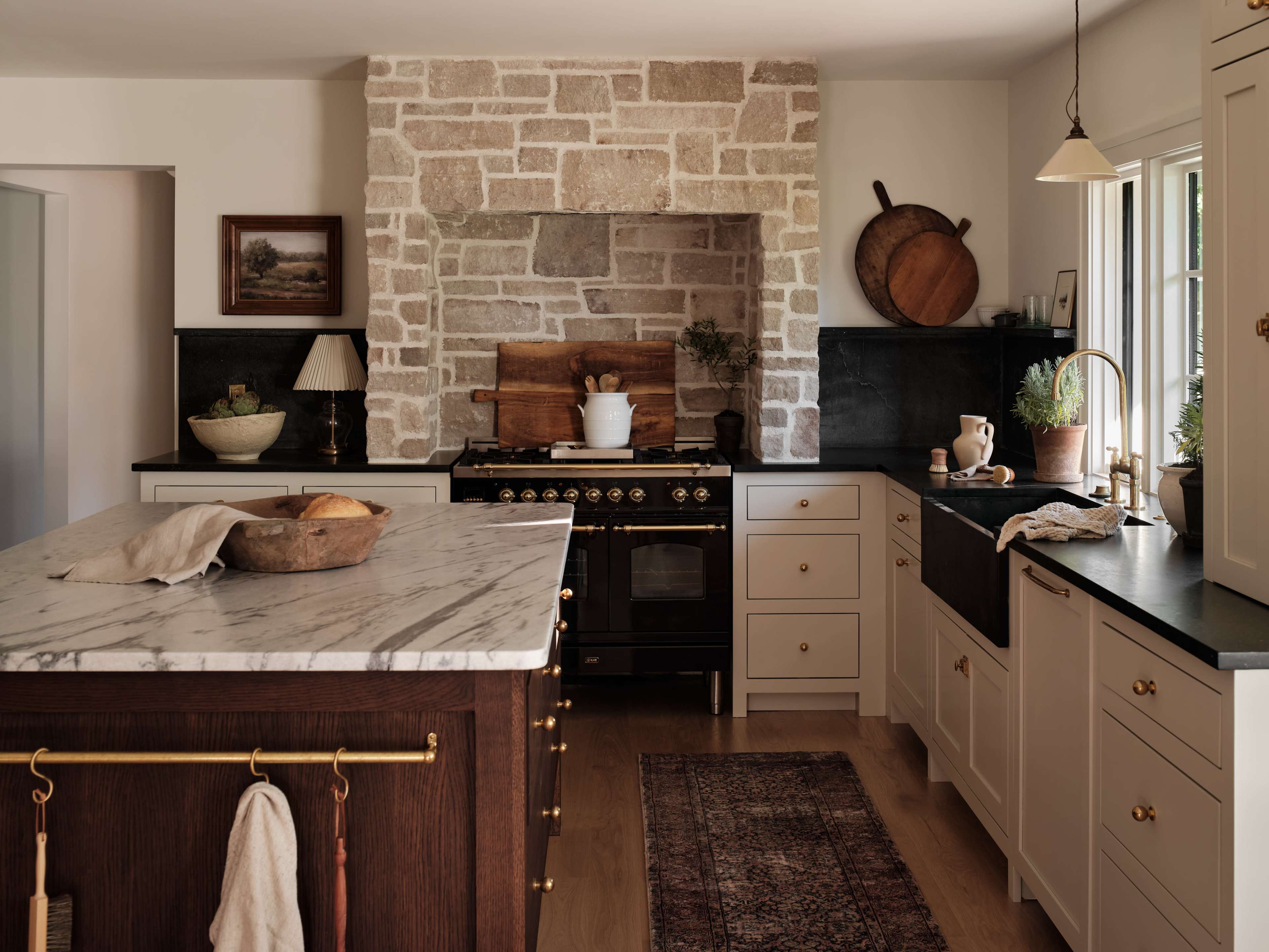 A modern kitchen features a large marble island, stone wall, and dark cabinetry with brass accents.