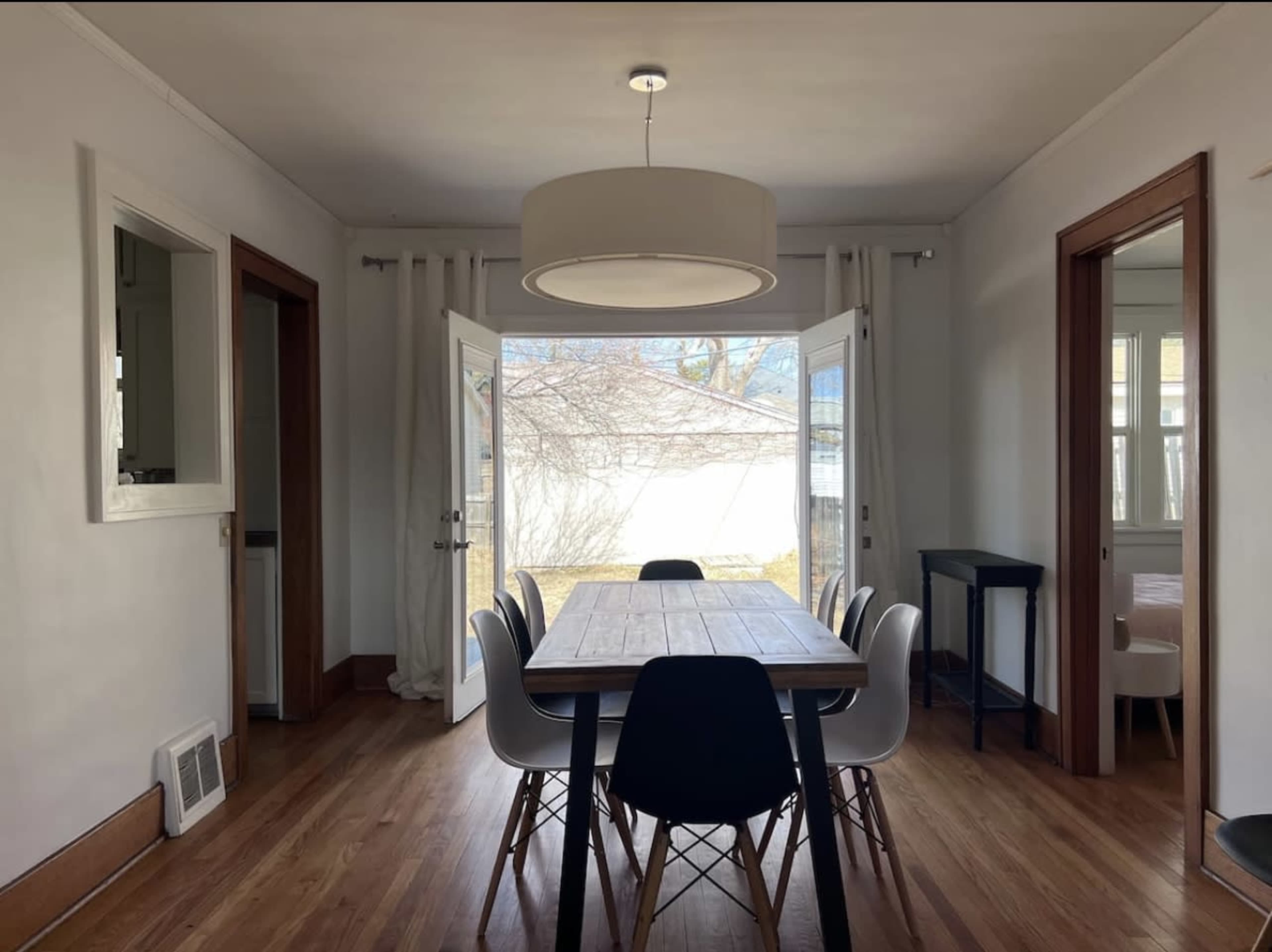 A dining area features a wooden table surrounded by chairs, with large windows and open doors leading to an outdoor space.