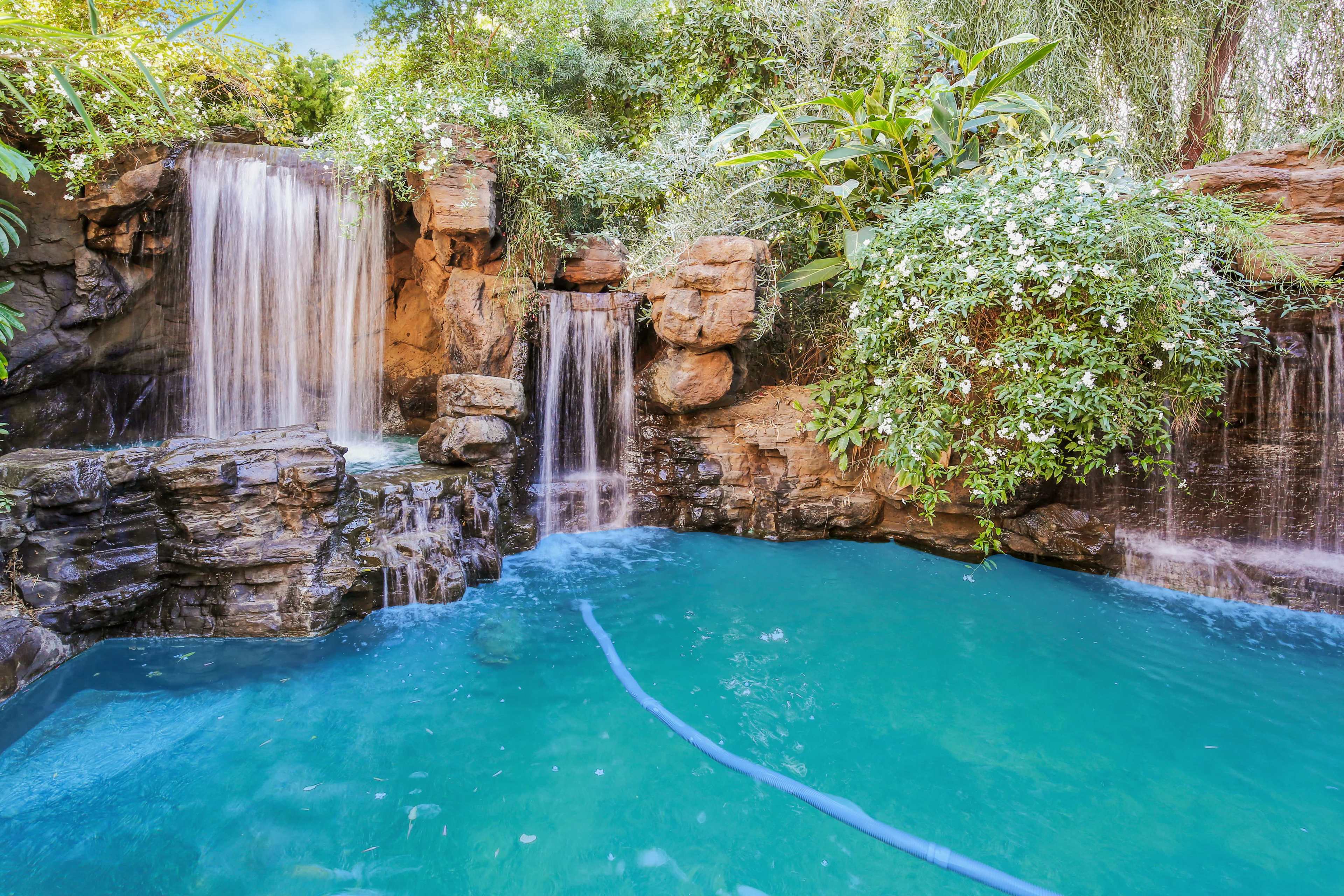 A lush garden with two waterfalls cascading over rocky formations into a turquoise pool.