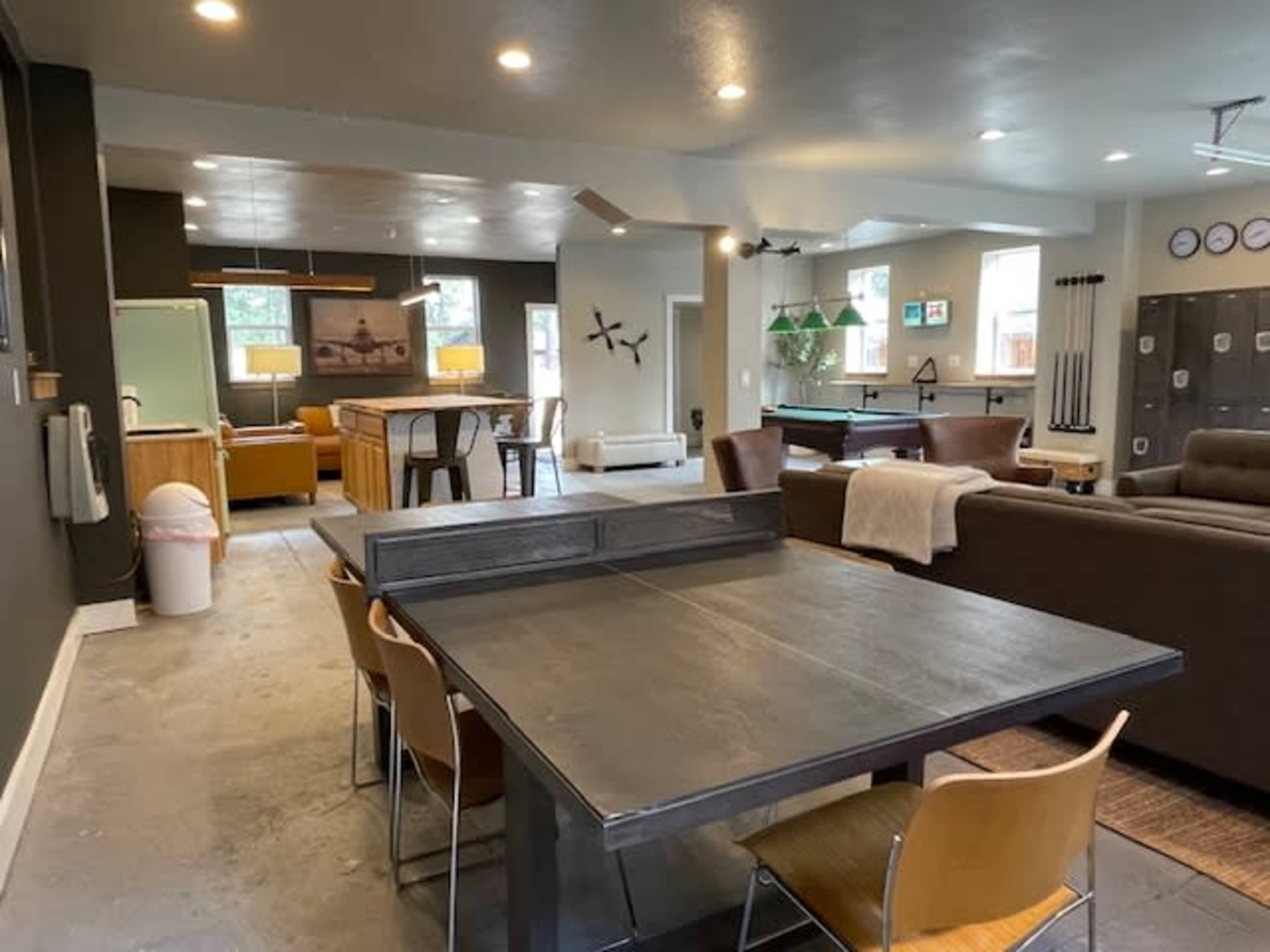 A spacious modern interior featuring a dining area with a metal table, adjacent living space with a sofa, a pool table, and various furniture pieces in a well-lit room.
