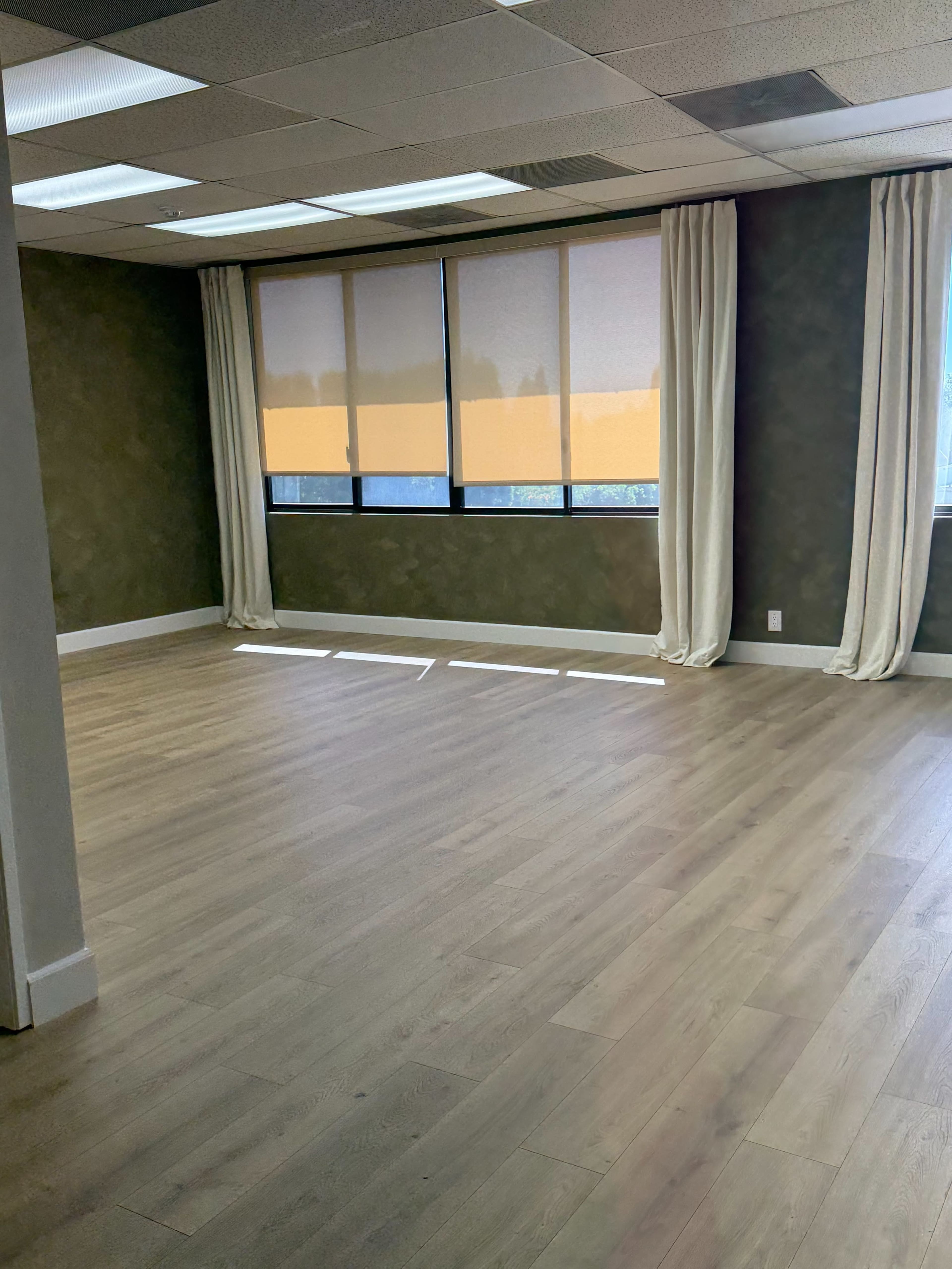 The image shows an empty room with wooden flooring, large windows covered by curtains, and a neutral-colored wall.