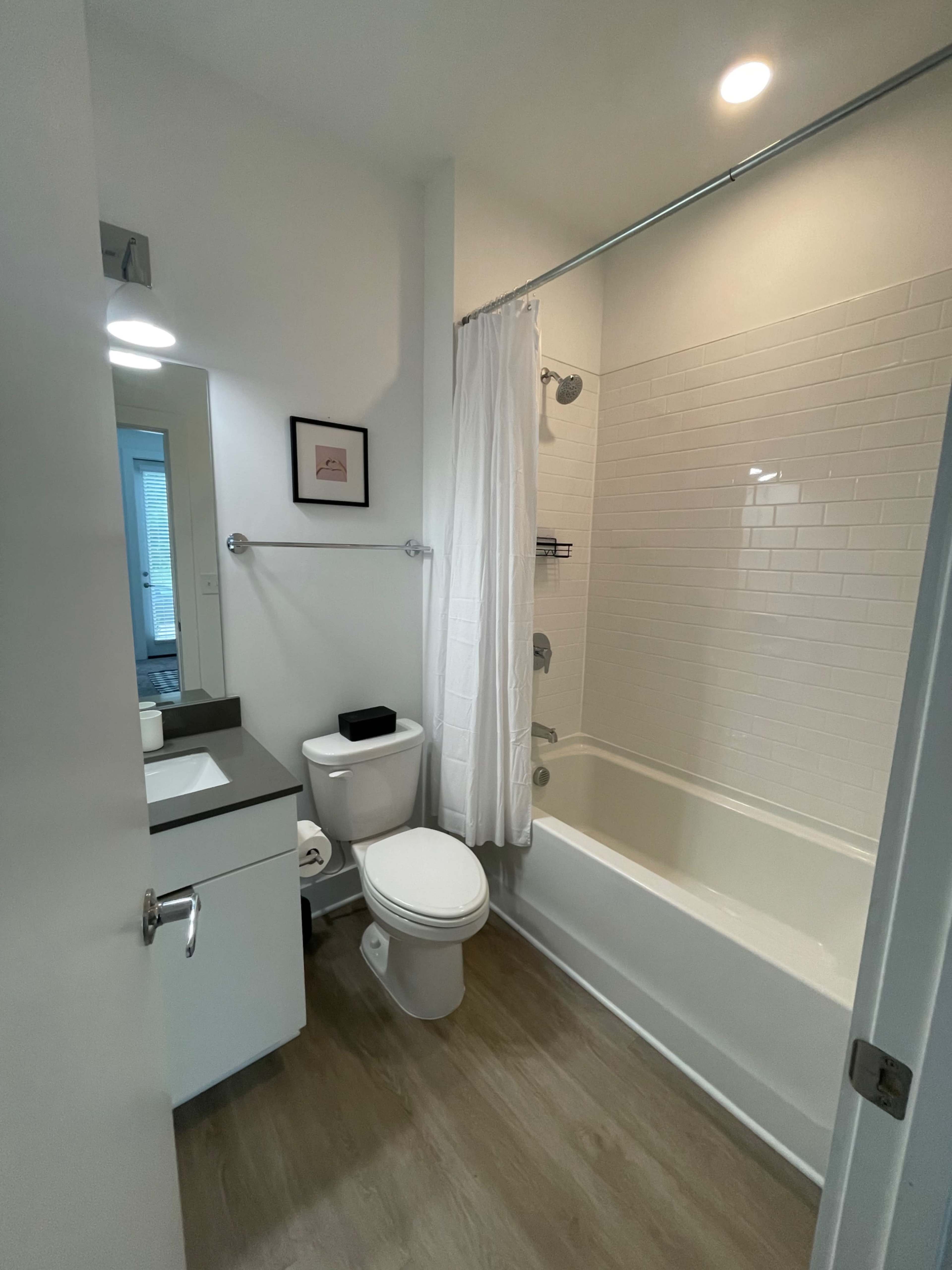 The image shows a modern bathroom featuring a bathtub combined with a shower, a toilet, and a sink with a mirror above it.