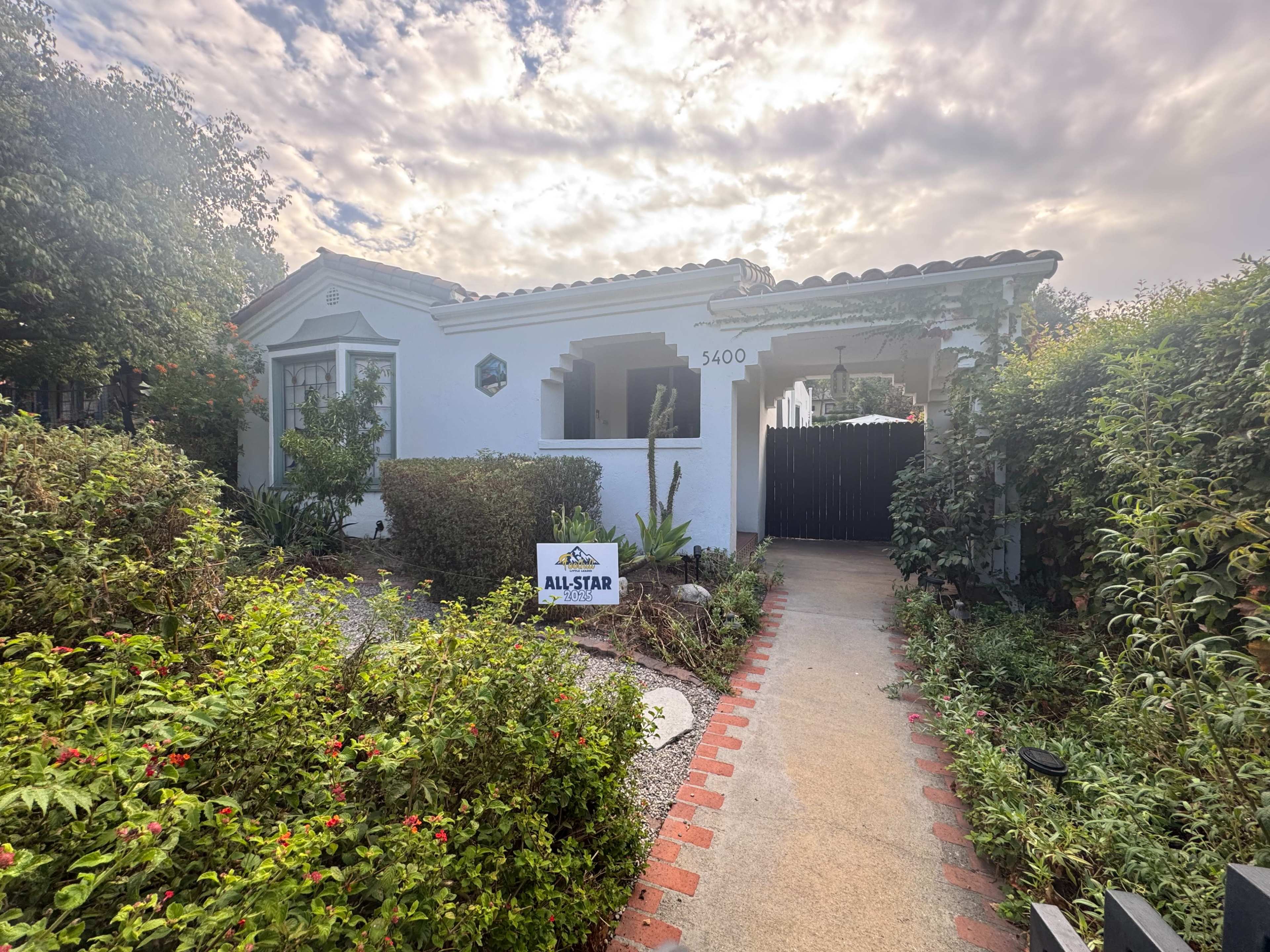 A single-story house with a white exterior and a clay tile roof is surrounded by greenery and has a "For Sale" sign in the front yard.