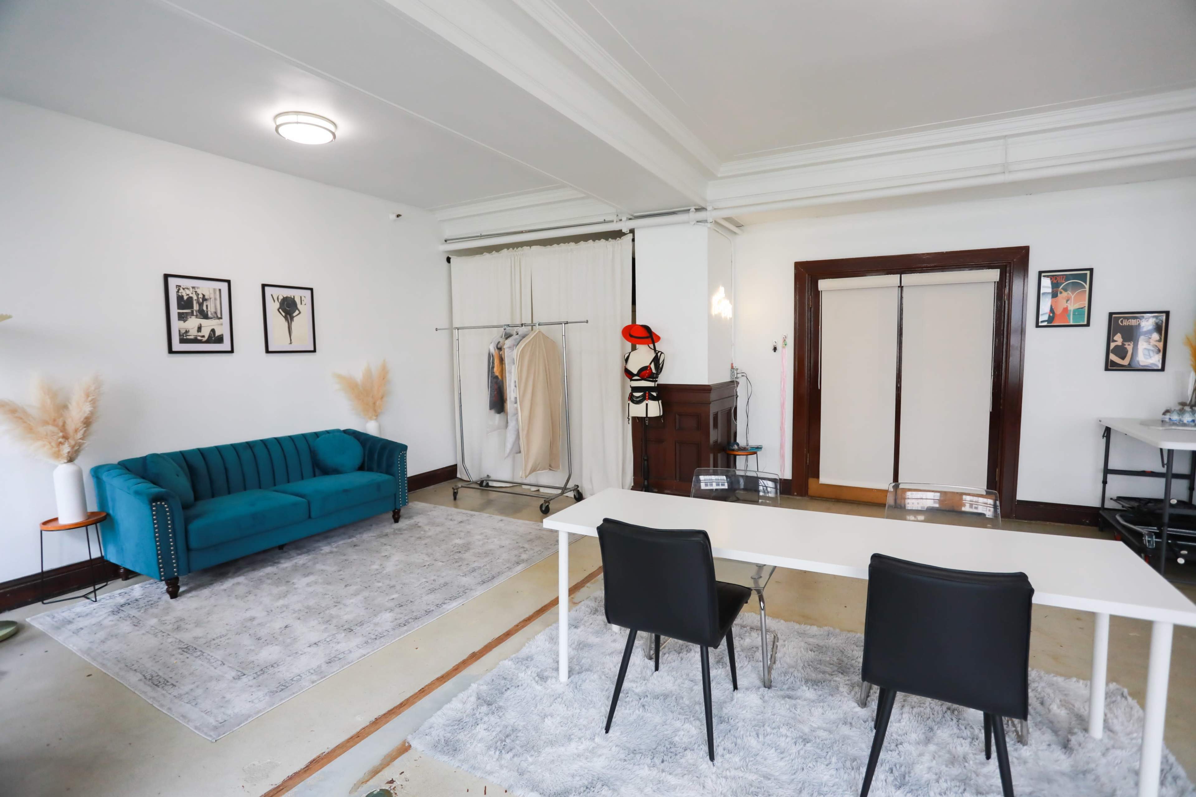 A well-lit room featuring a teal velvet sofa, a clothing rack with garments, a white table with two black chairs, and decorative elements like pampas grass.