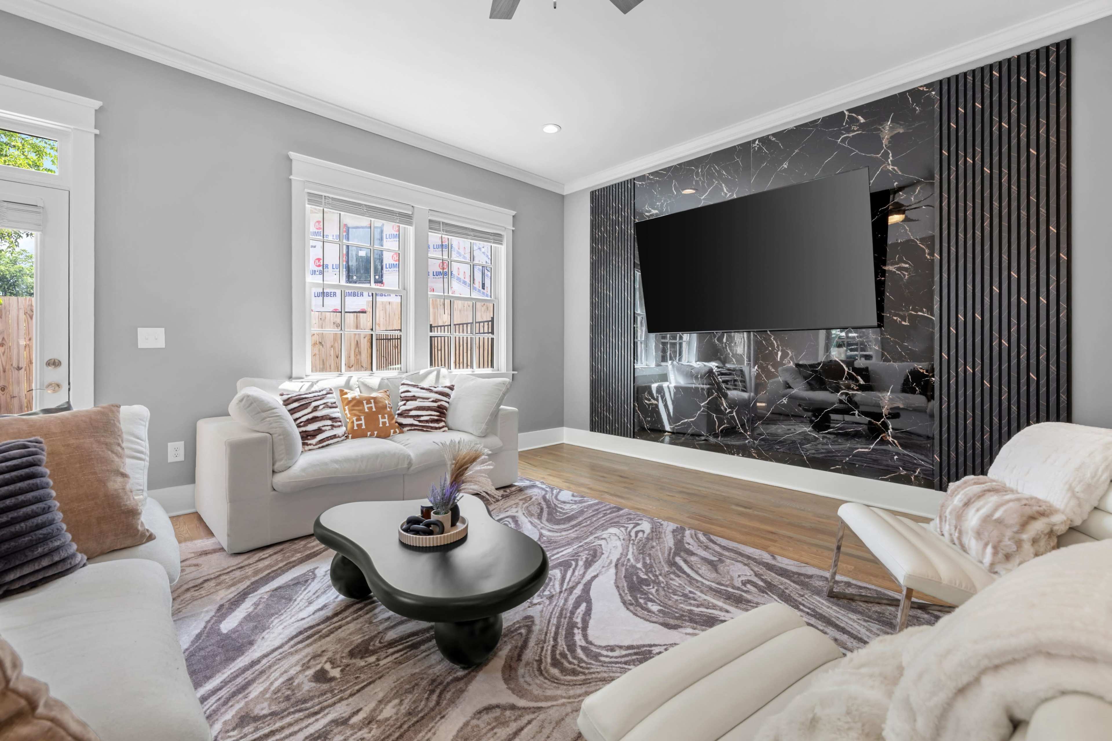 The image shows a modern living room featuring a white sofa, a dark coffee table, and a wall-mounted television above a glossy black decorative wall, with large windows allowing natural light to enter.