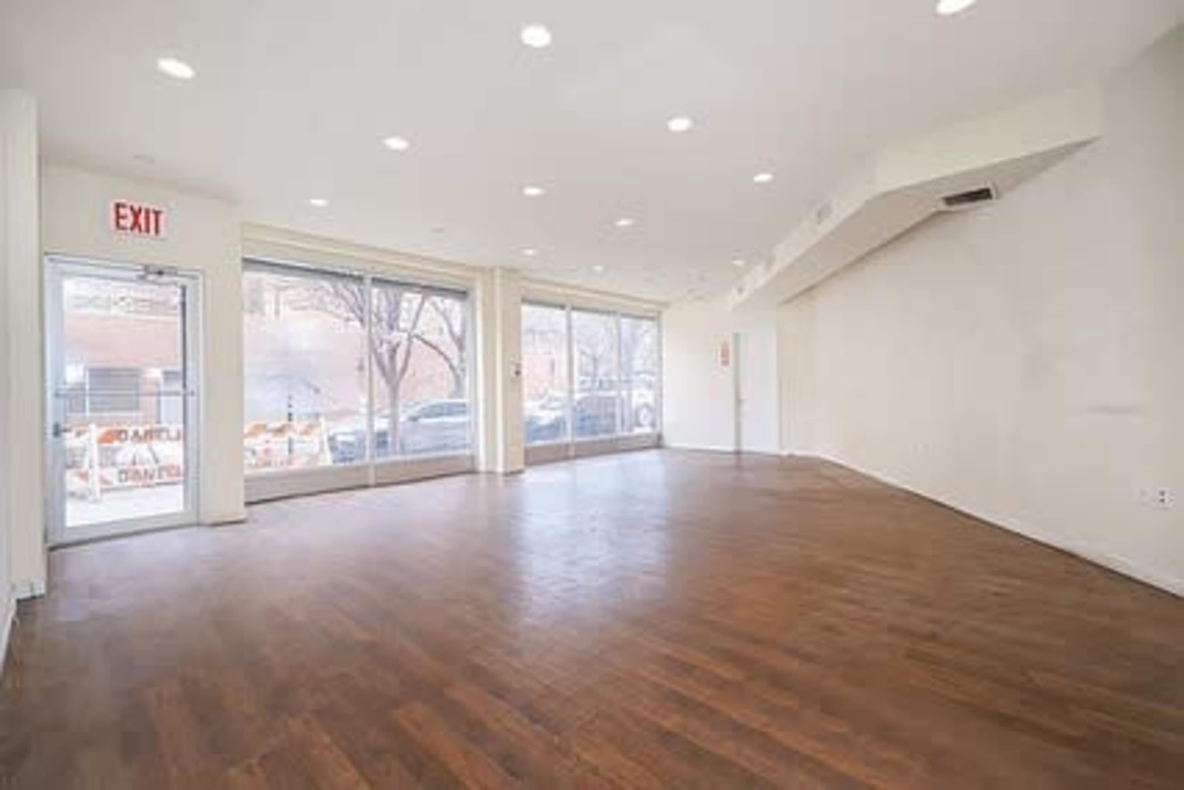 An empty retail space with hardwood flooring, large windows, and an exit sign near the entrance.
