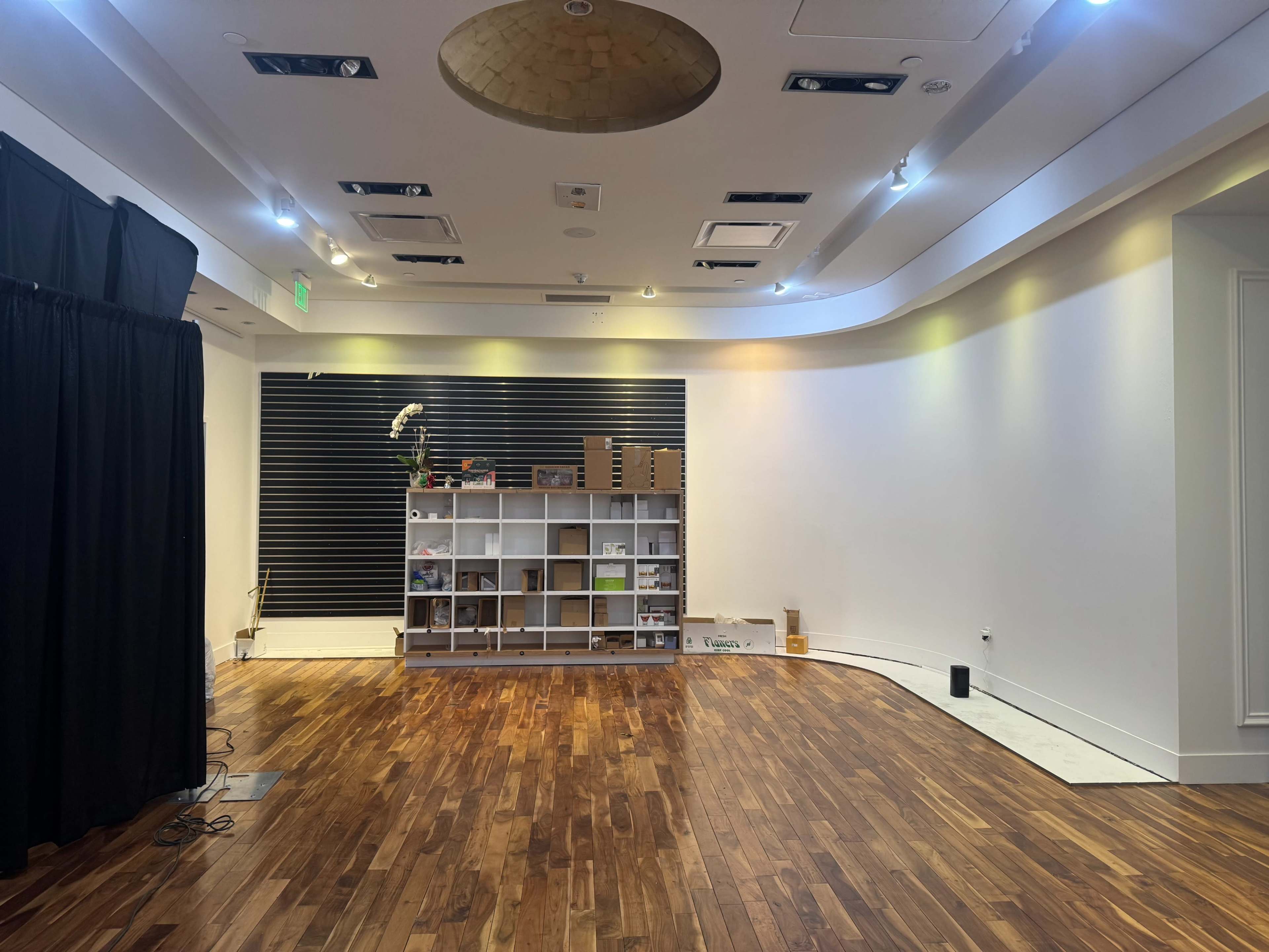 The image shows an empty room with wooden flooring, featuring a white shelving unit against a wall and a black curtain on one side.