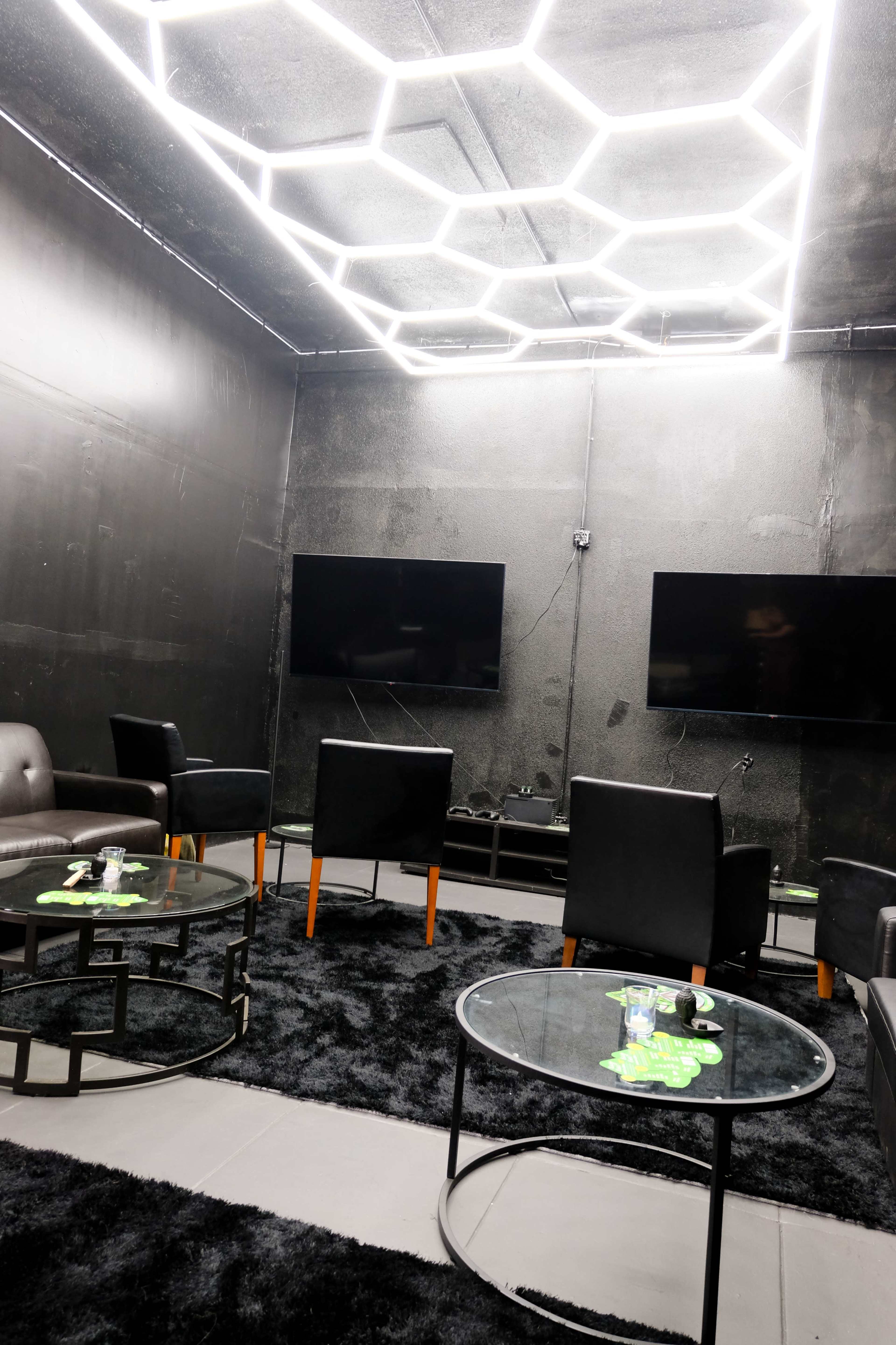 The room features black walls, a hexagonal light fixture, two televisions, and lounge chairs arranged around glass tables on a dark carpet.