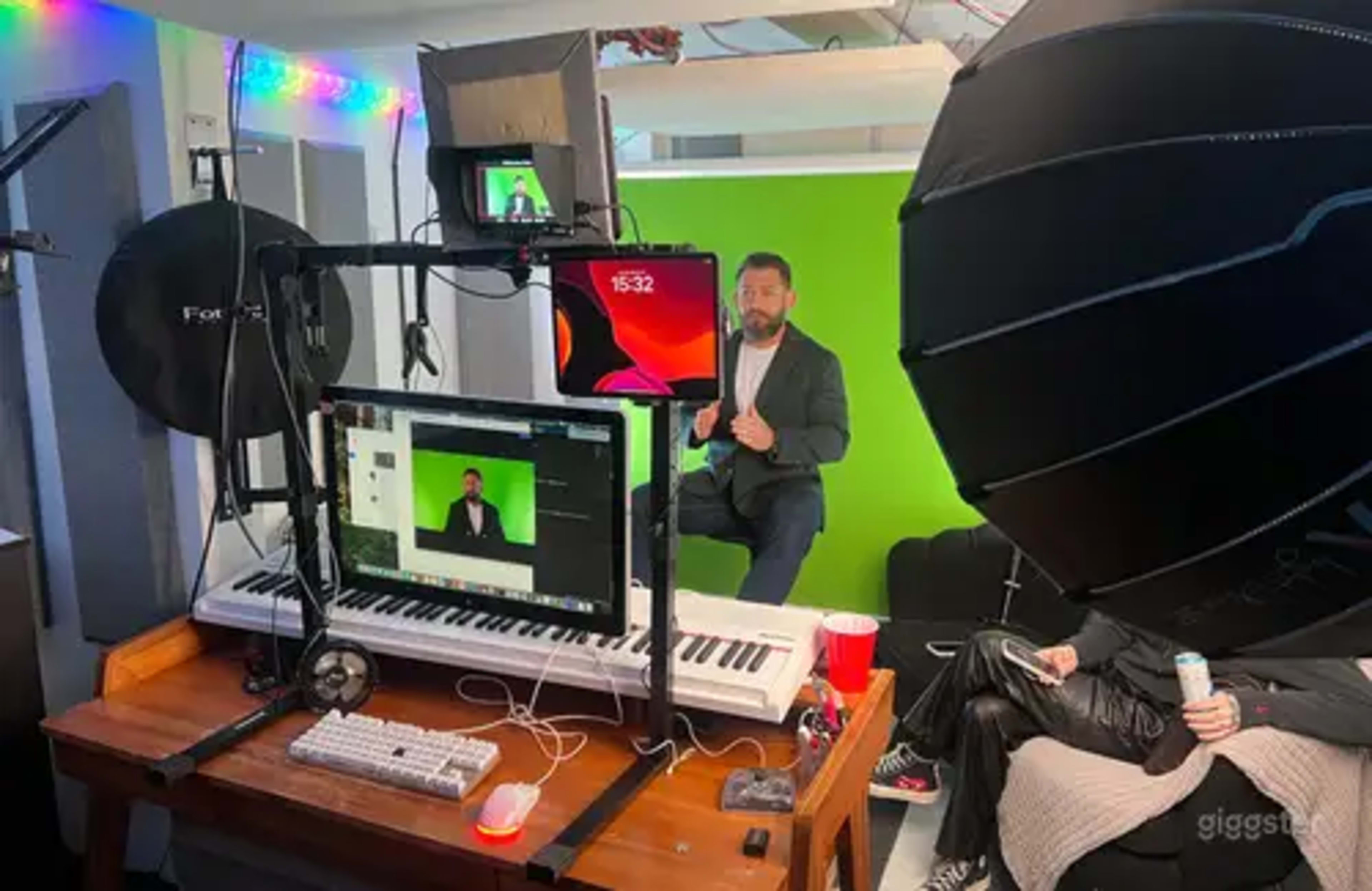 A video recording setup features a man in a suit sitting in front of a green backdrop, surrounded by lighting equipment, a computer, and a keyboard.