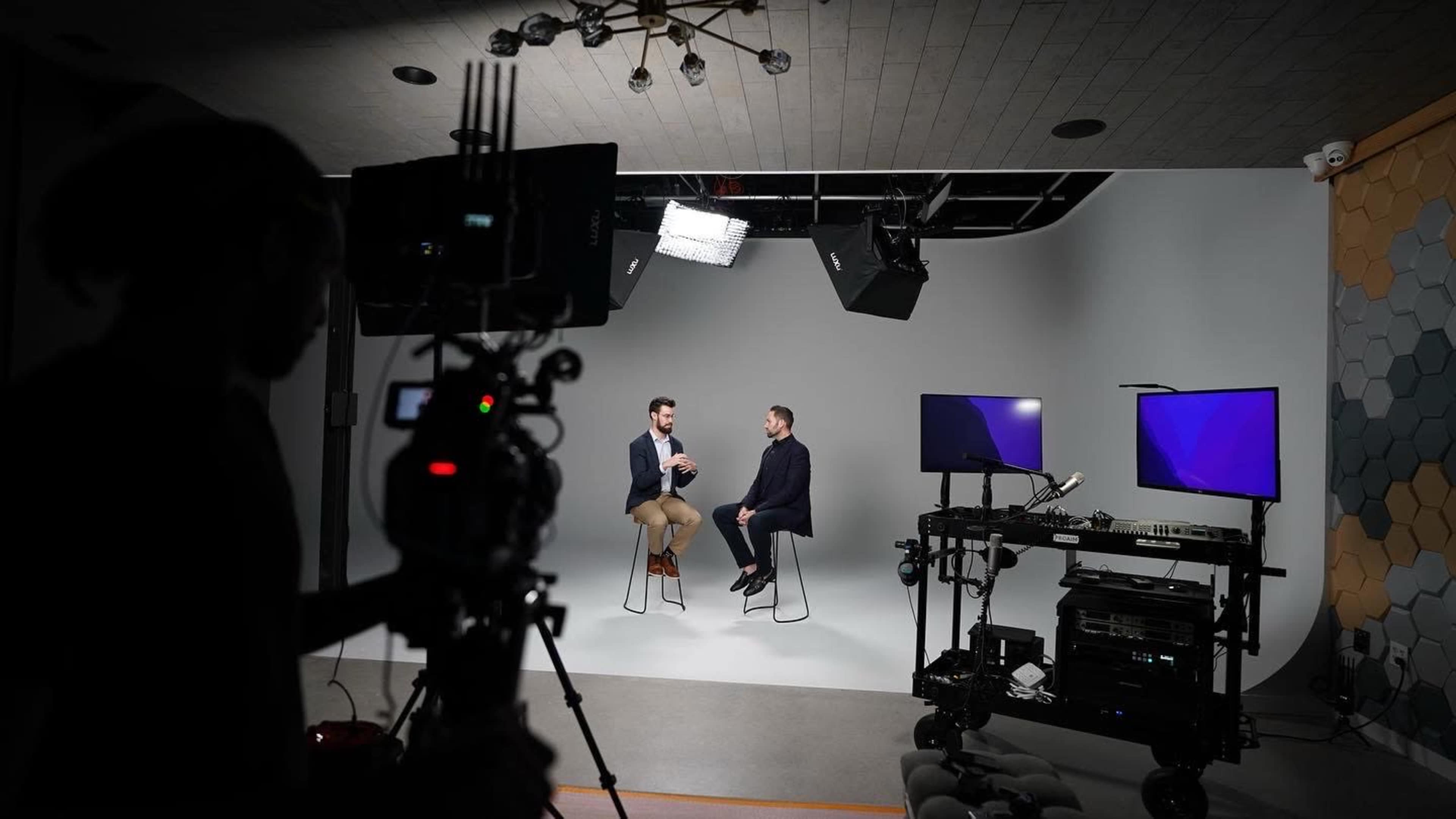 Two men are seated on stools in a studio setting, engaged in conversation while a camera and lighting equipment are positioned nearby.