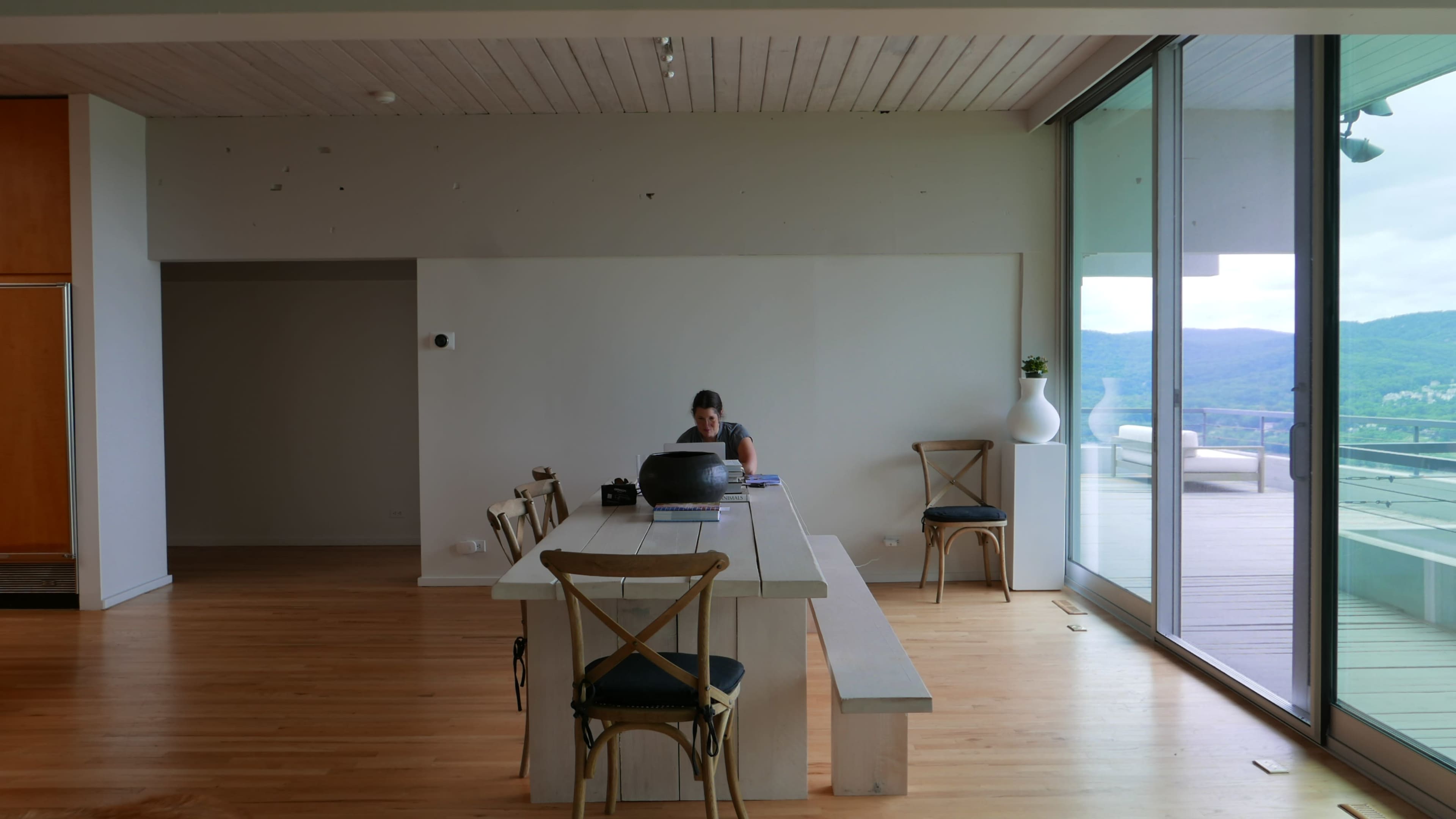 A person is working on a laptop at a long wooden table in a modern, minimalist room with large glass doors leading to a balcony.
