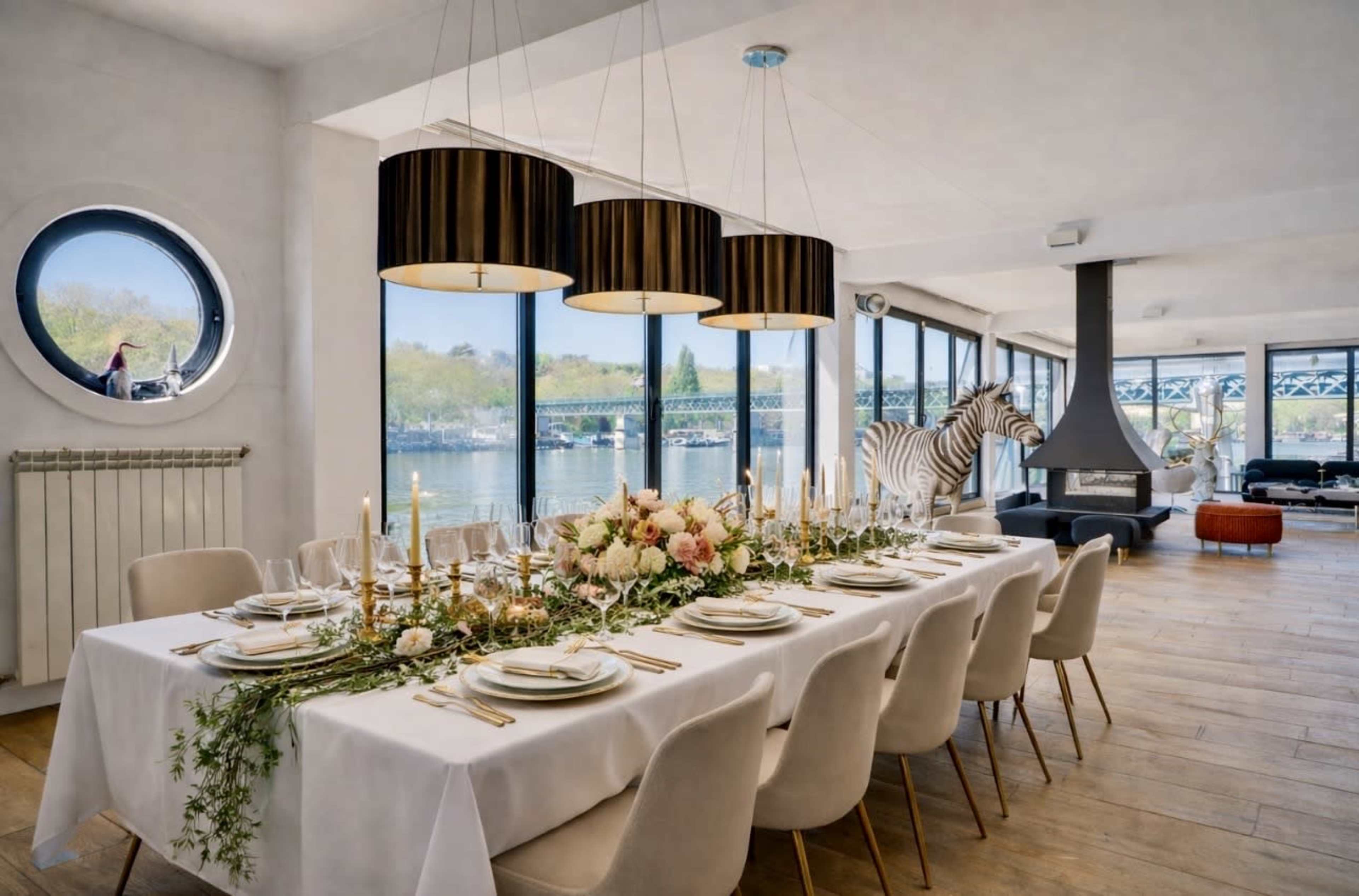 A spacious dining area features a long table set with elegant dishes and candle holders, flanked by large windows with a view of the water and a zebra figurine in the background.