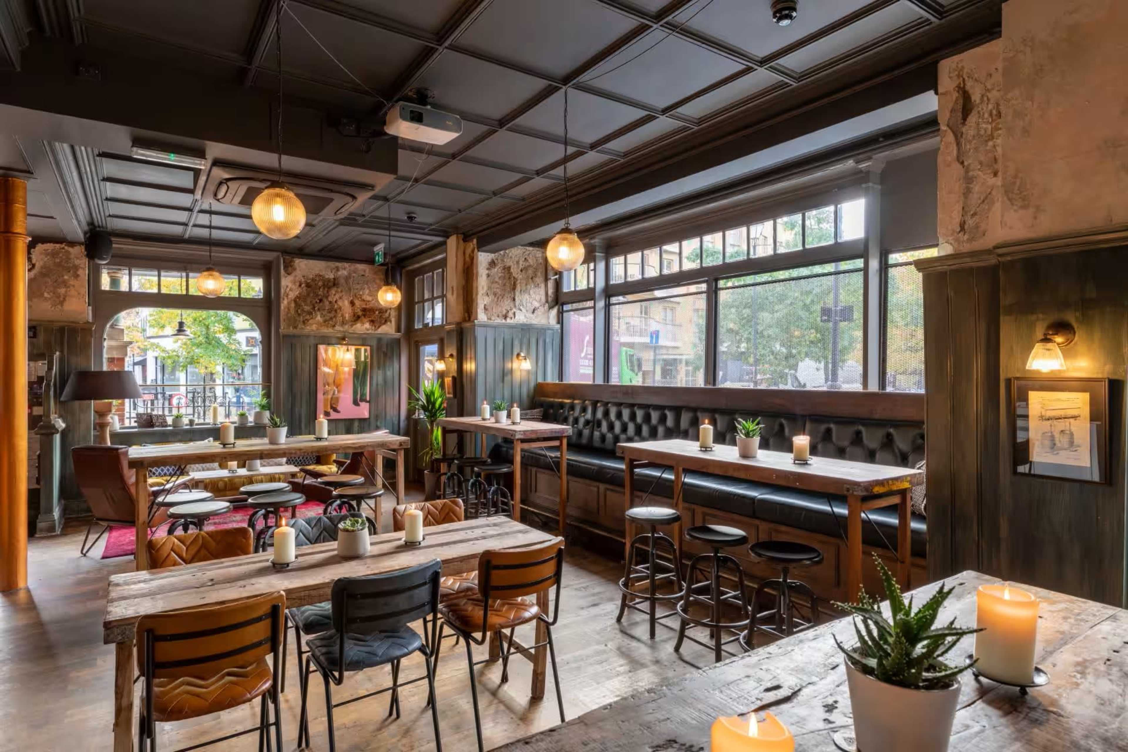 The image shows an interior of a cozy restaurant with wooden tables, seating areas, and large windows allowing natural light to enter.