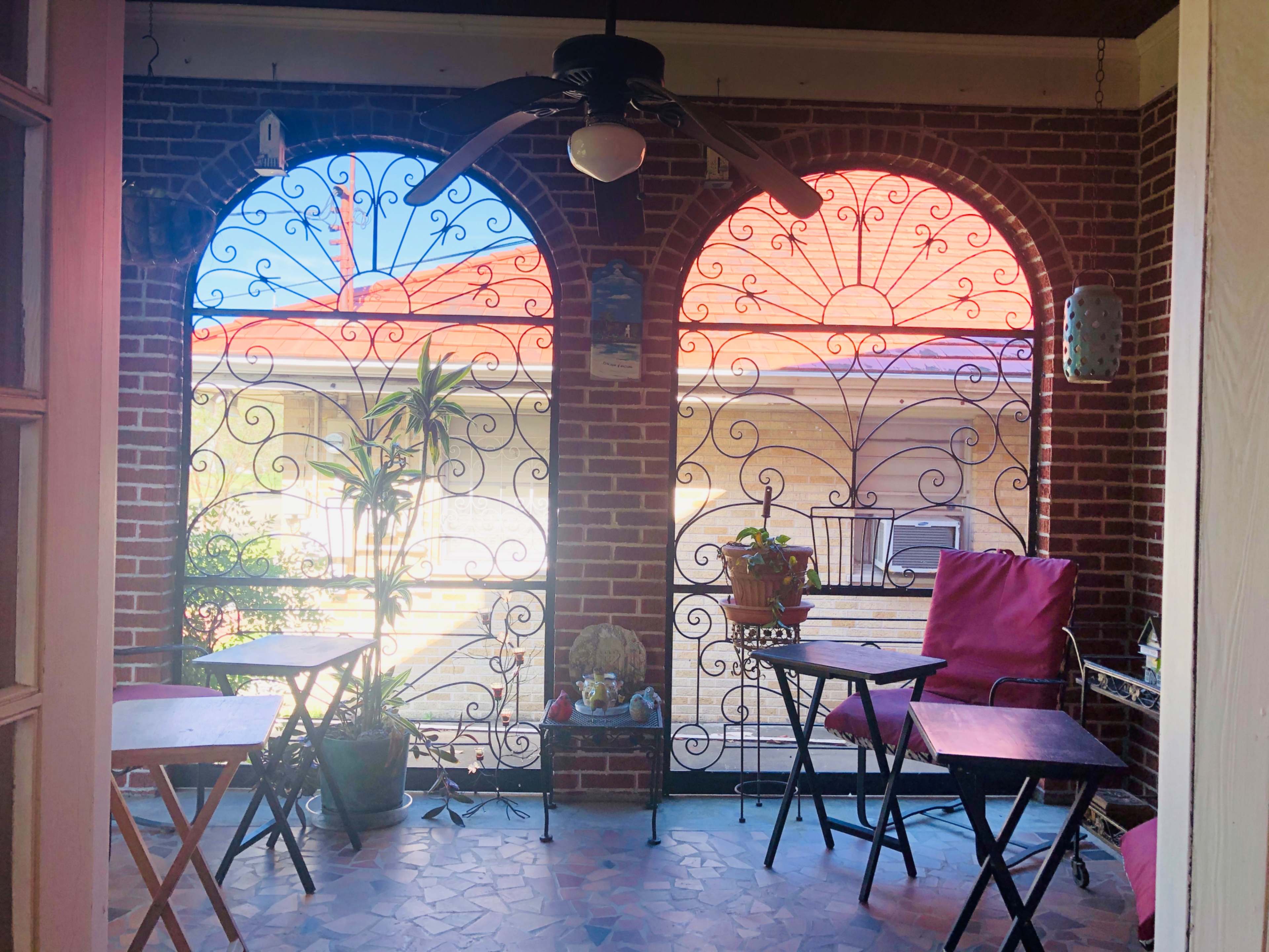 The image shows a brick porch with two arched openings, each featuring decorative wrought iron work, and wooden tables with chairs placed on a tiled floor.