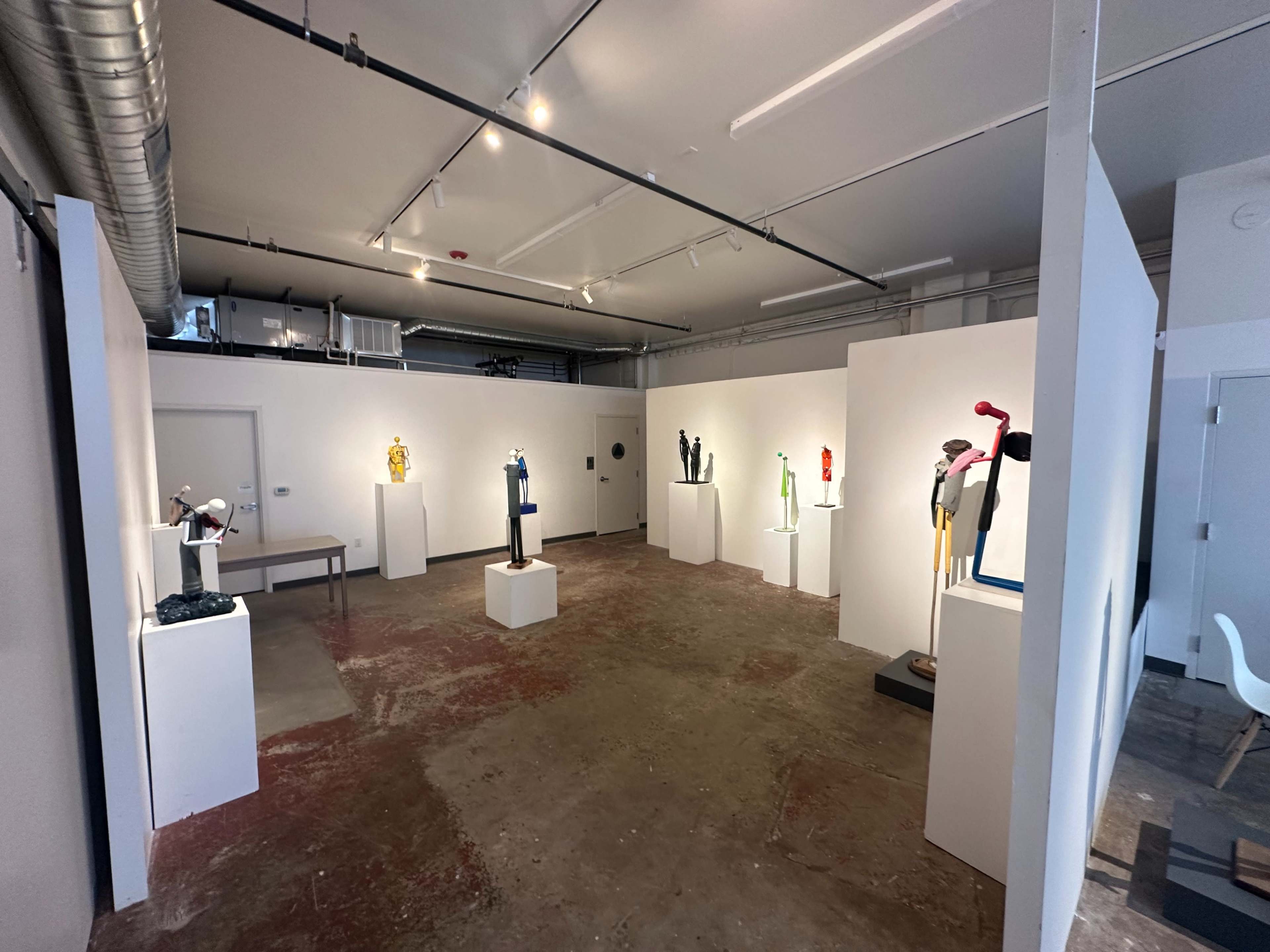 An art gallery featuring white pedestals displaying various sculptures against a backdrop of gray walls and a concrete floor.