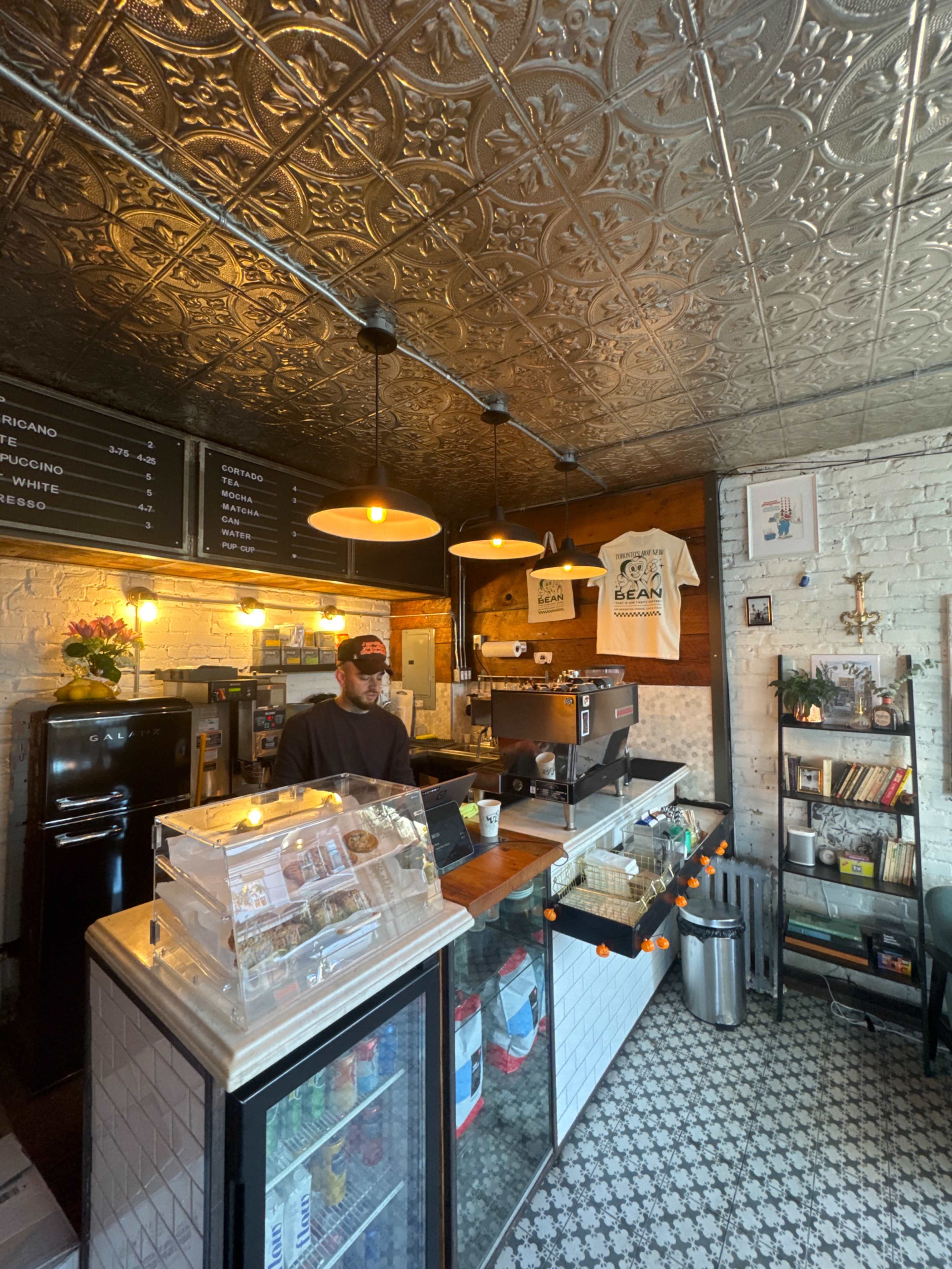 A barista stands behind the counter of a cozy café featuring vintage decor and menu listings on the wall.