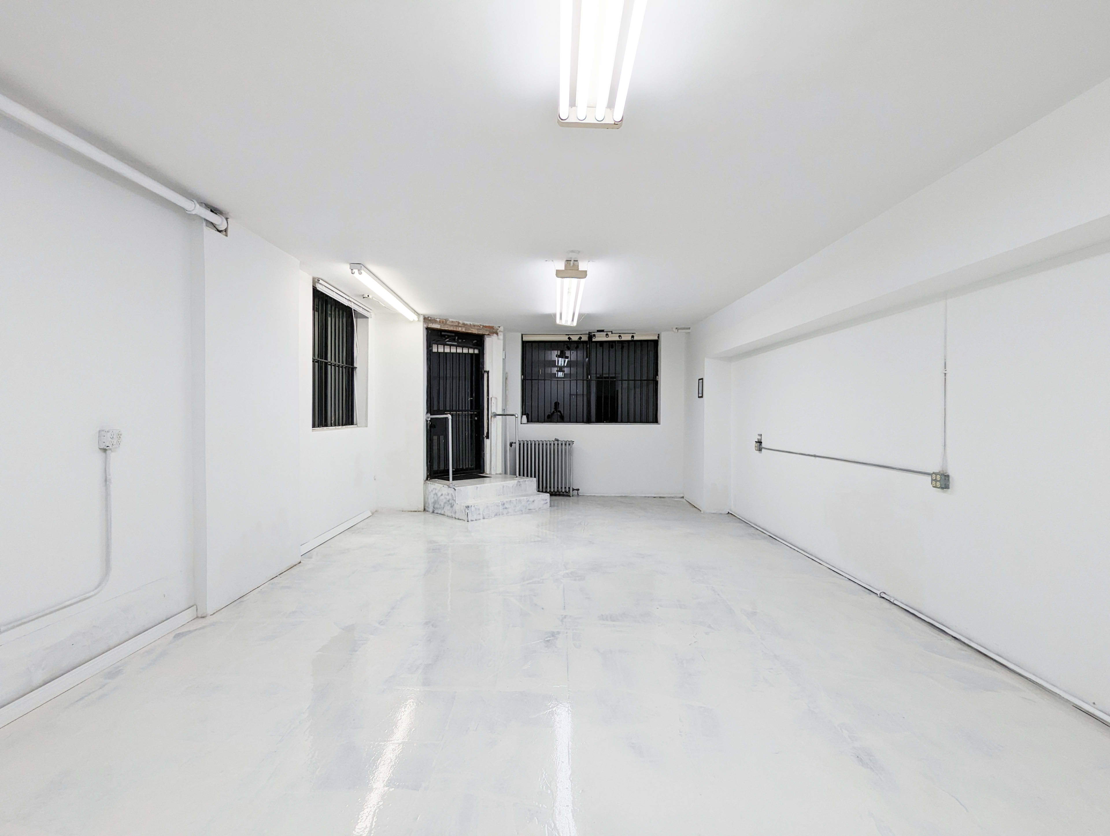 The image shows a spacious, empty room with white walls and a polished floor, illuminated by overhead fluorescent lights.