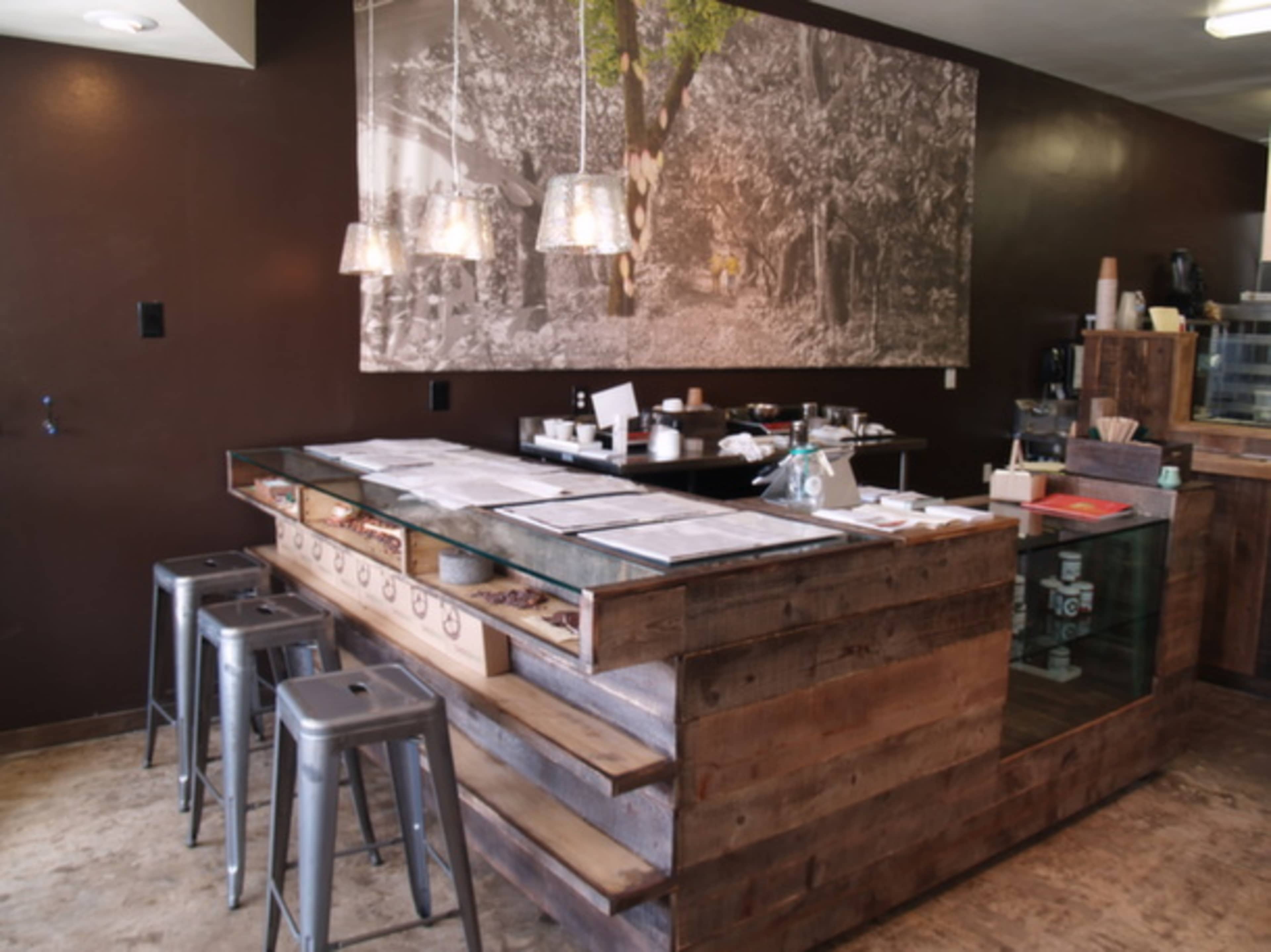 A coffee shop interior features a wooden counter with metal stools and a large mural of a tree on the wall.