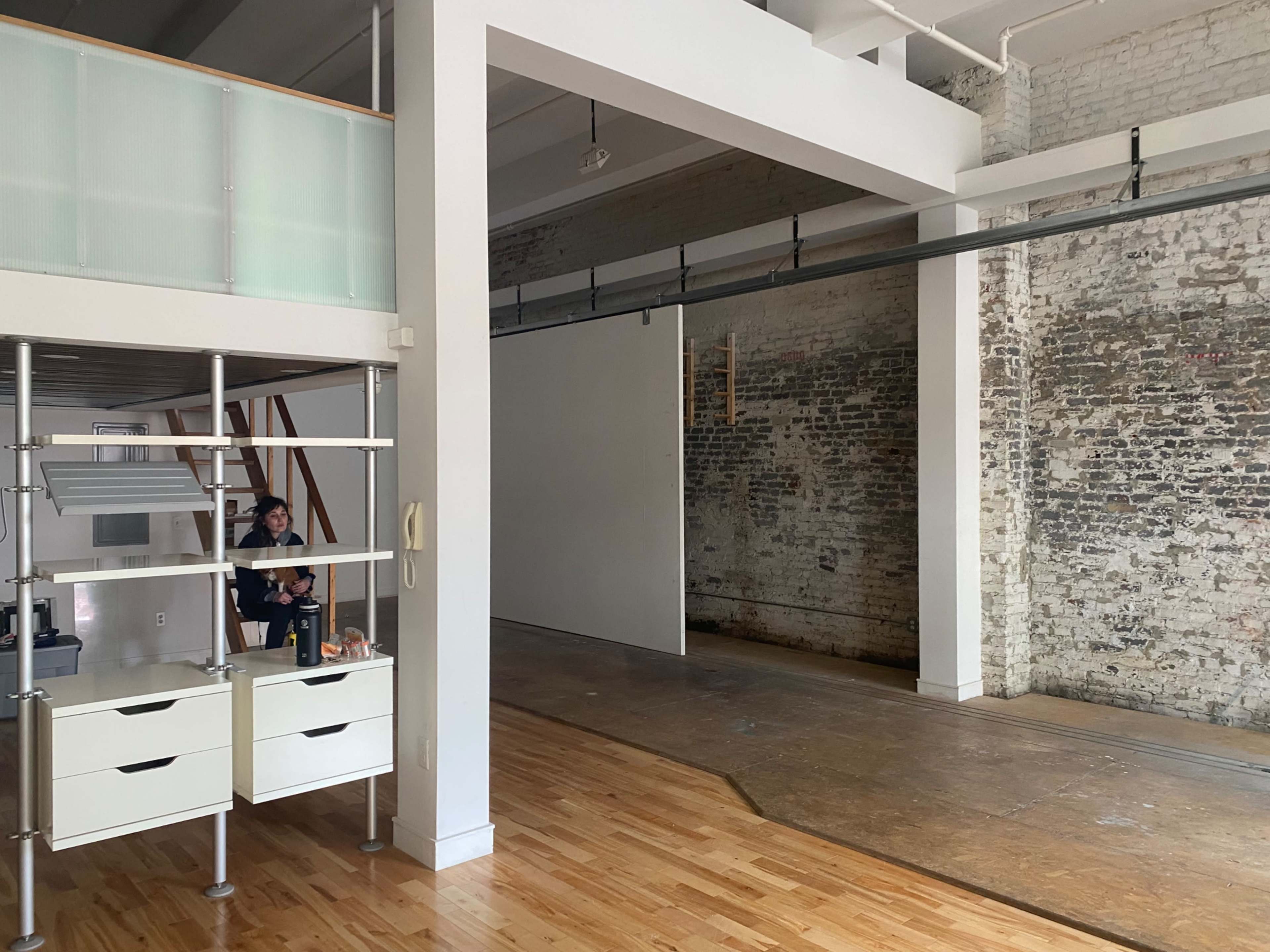 The image shows a spacious room with exposed brick walls, hardwood floors, and a minimalist office setup featuring a shelving unit and some furniture.