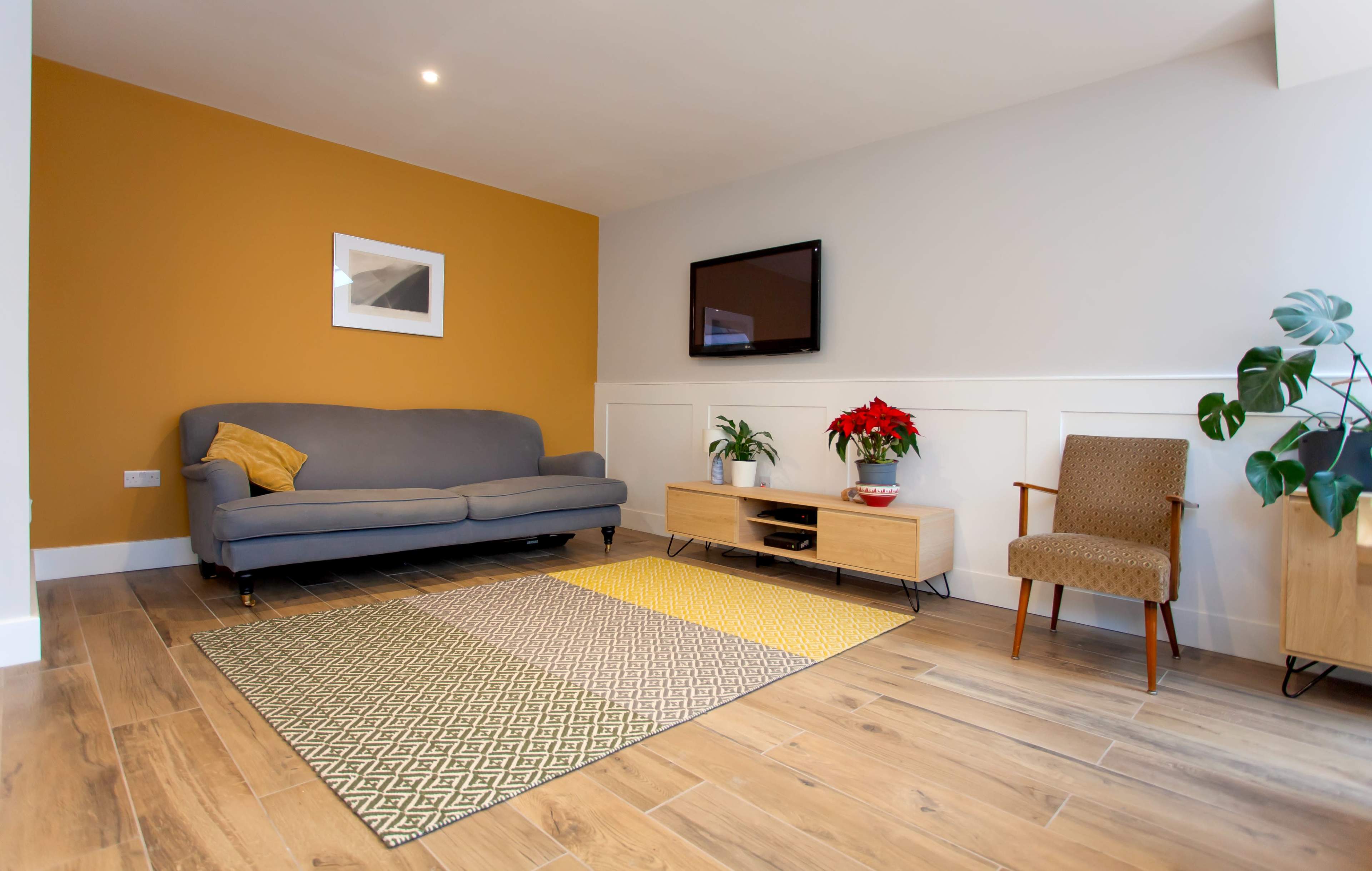The image shows a modern living room with a gray sofa, a wooden media unit, a TV mounted on the wall, and a patterned rug on the floor.
