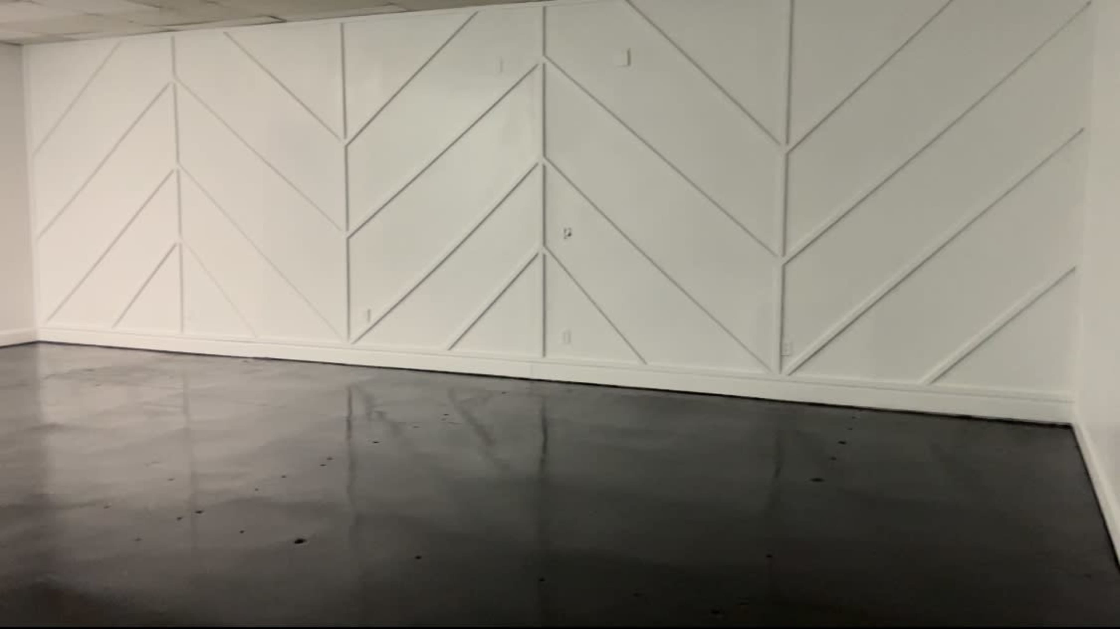 The image shows a sparsely furnished room with a polished concrete floor and white chevron-patterned walls.