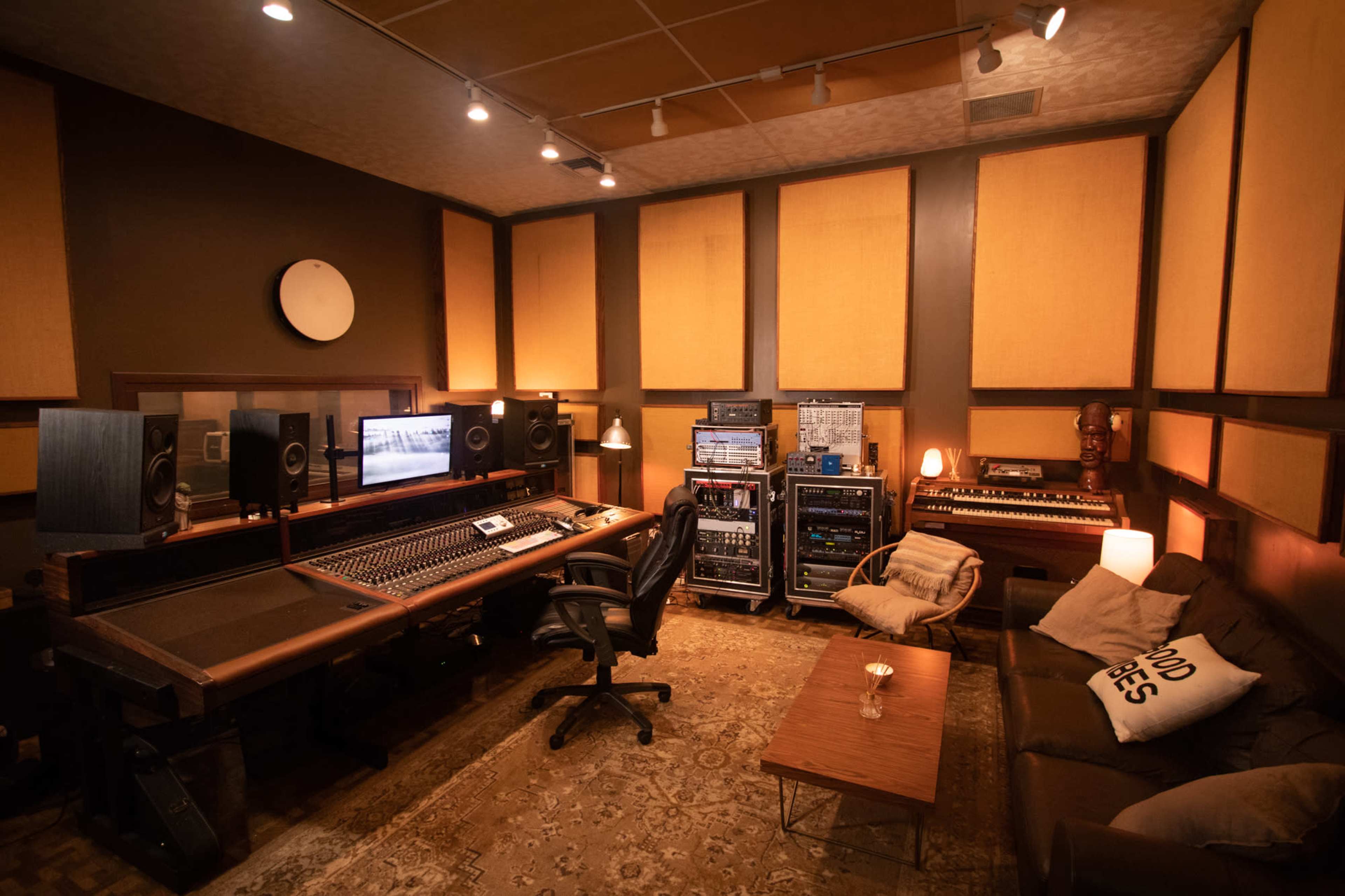 A recording studio with a large mixing console, audio equipment racks, and seating arranged around a central coffee table.
