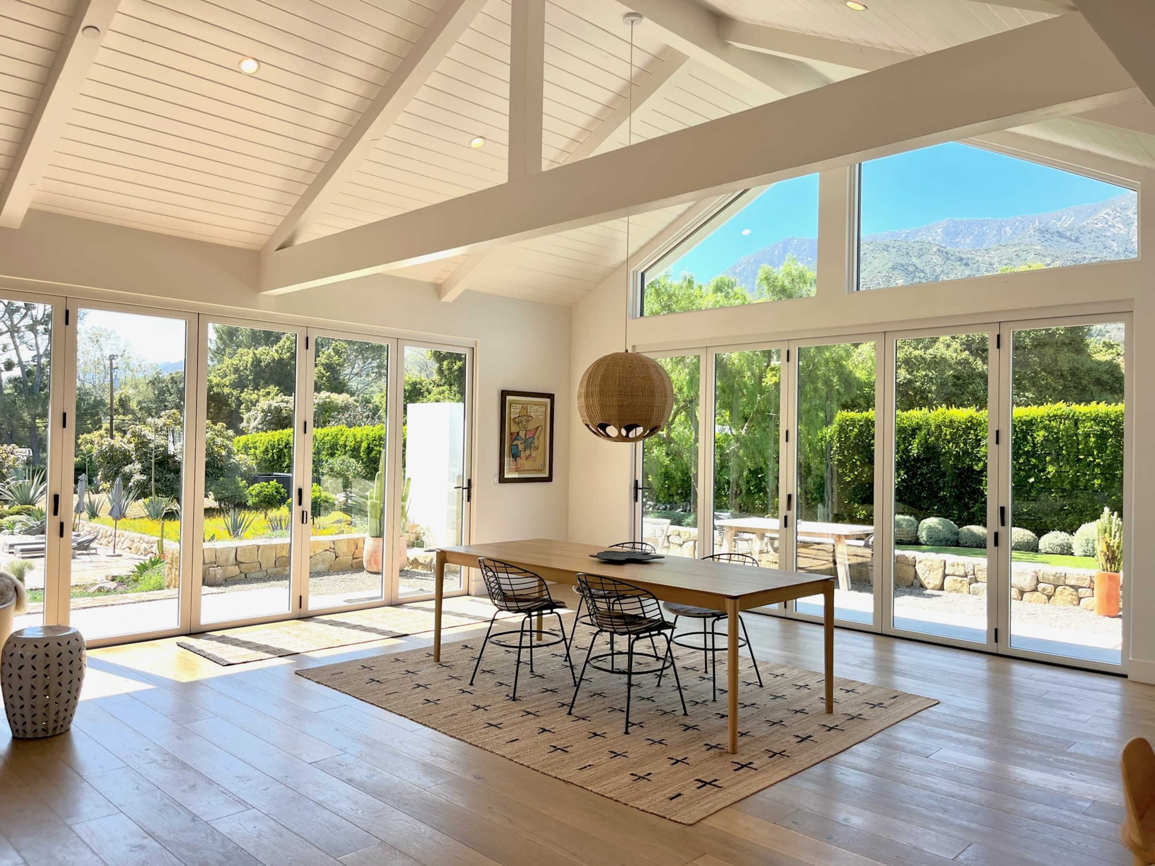 A bright, airy dining area features a wooden table surrounded by black chairs, with large windows offering a view of a landscaped garden.
