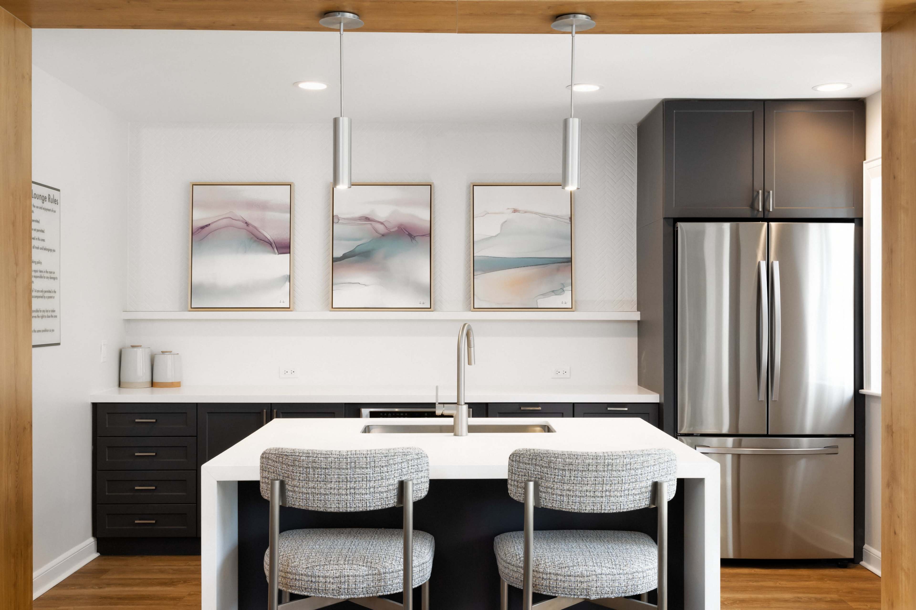 A modern kitchen features a central island with two bar stools, stainless steel appliances, and framed abstract art on the wall.