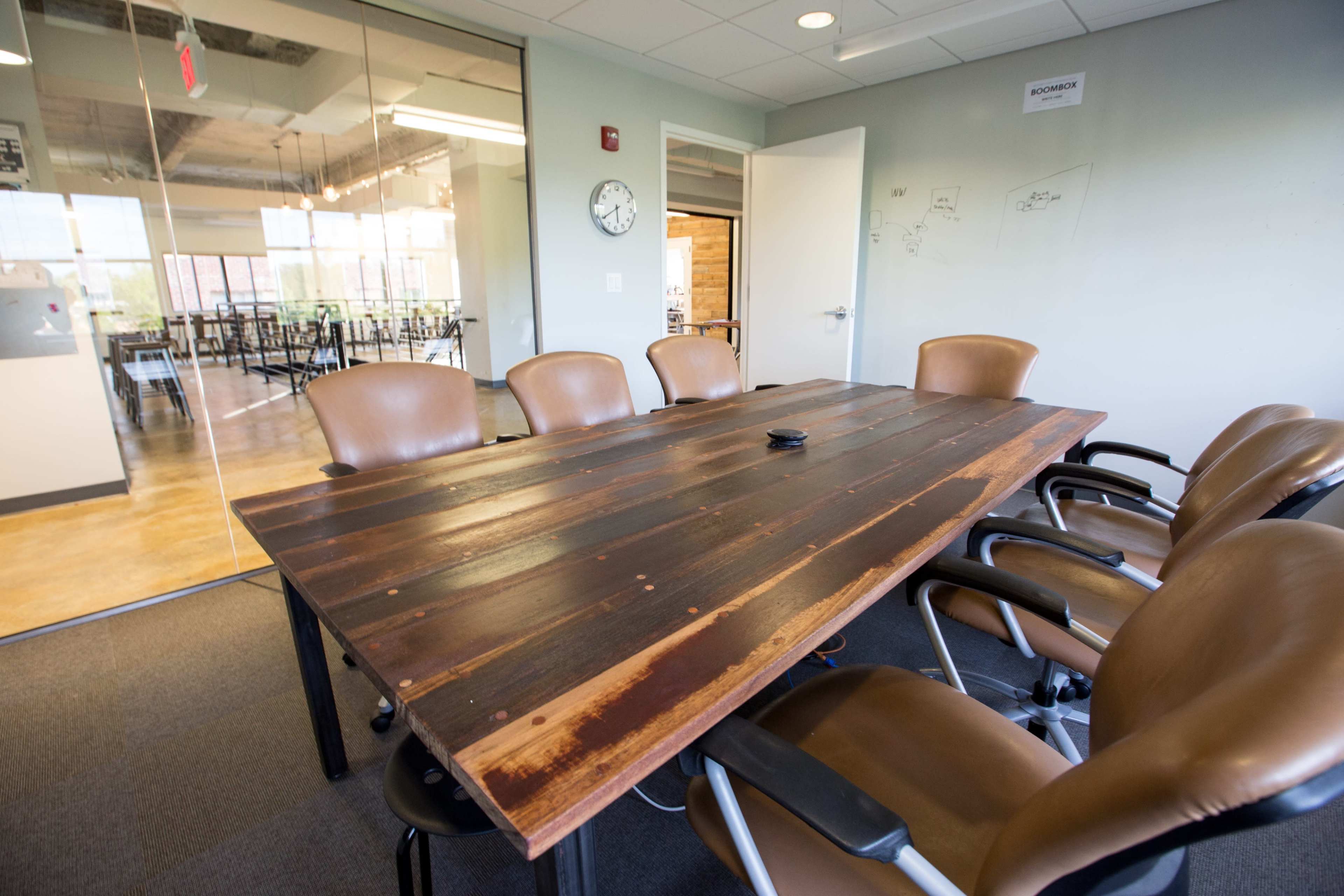 A wooden conference table with six chairs surrounds it in a meeting room featuring glass walls and a wall clock.