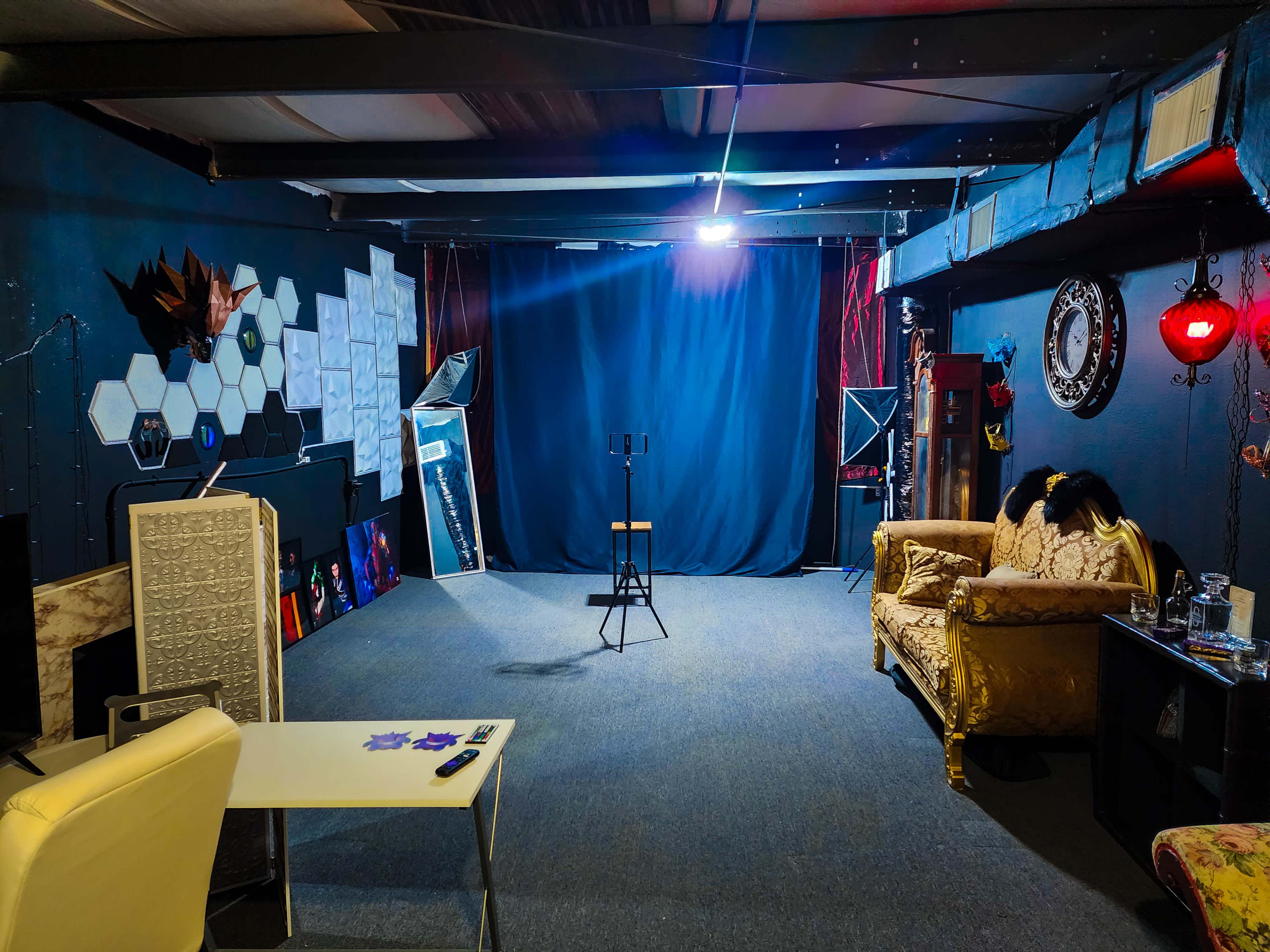 A dimly lit studio space featuring a black backdrop, a spotlight above a stool, and assorted furniture including a golden sofa and a white table.
