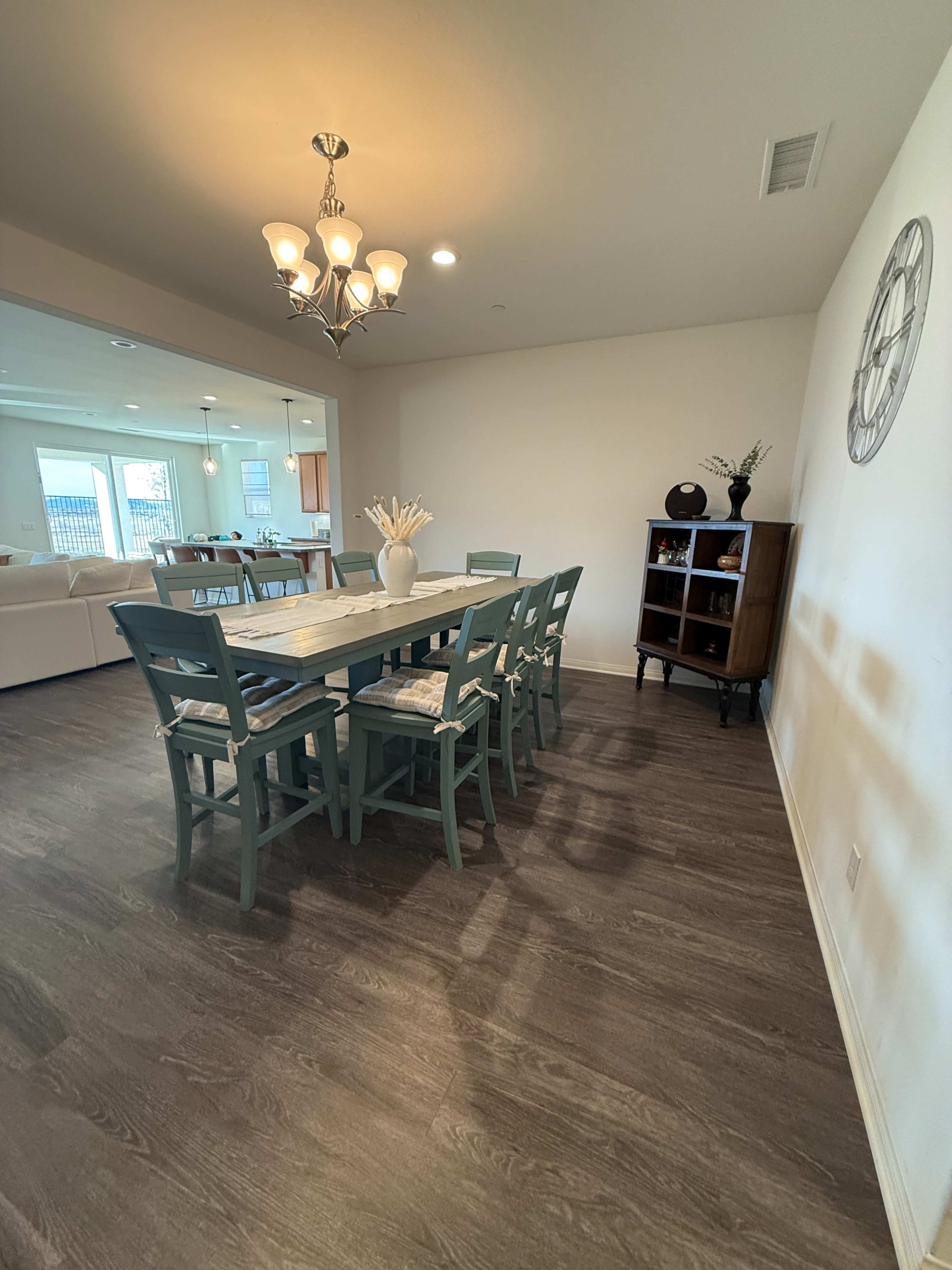 A dining area features a long wooden table surrounded by six chairs, with a chandelier above and a cabinet against the wall.