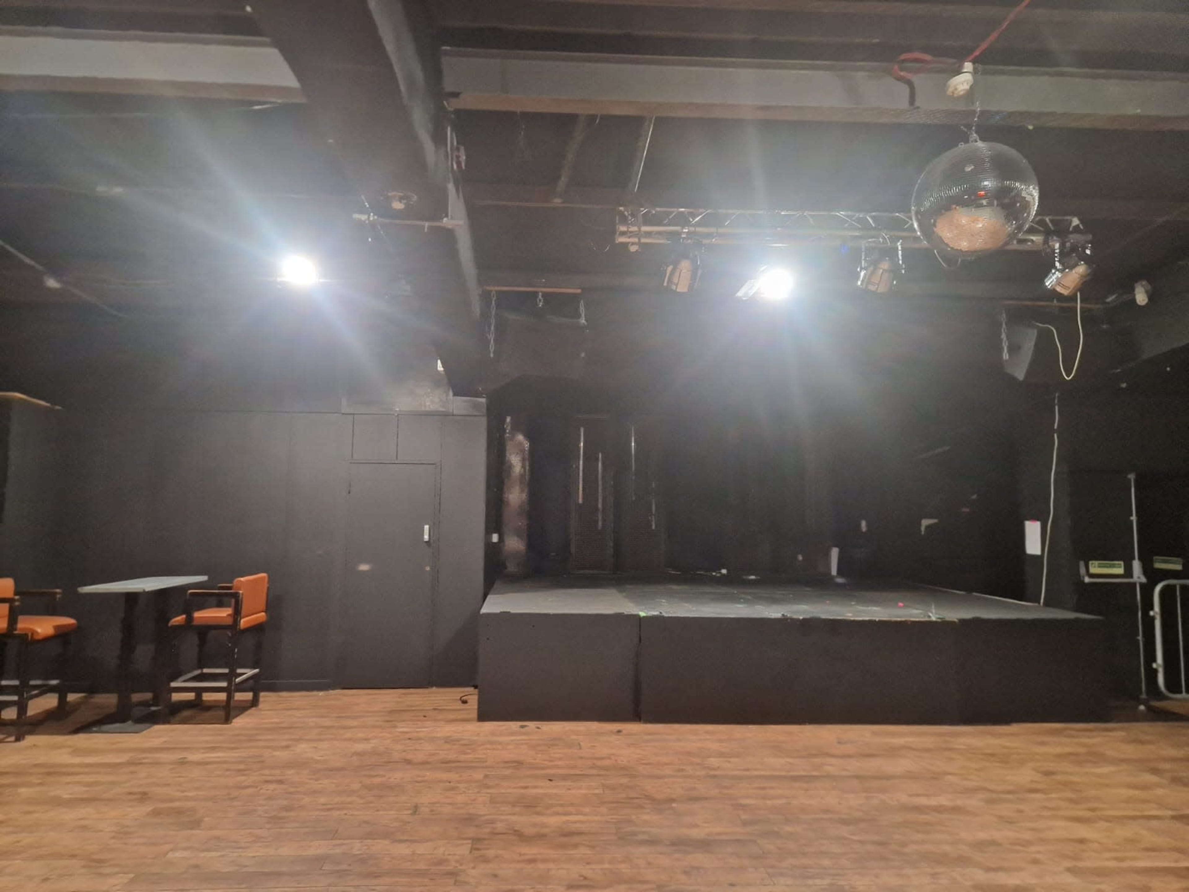 The image shows a dimly lit performance stage with a disco ball and a seating area featuring high tables and chairs.