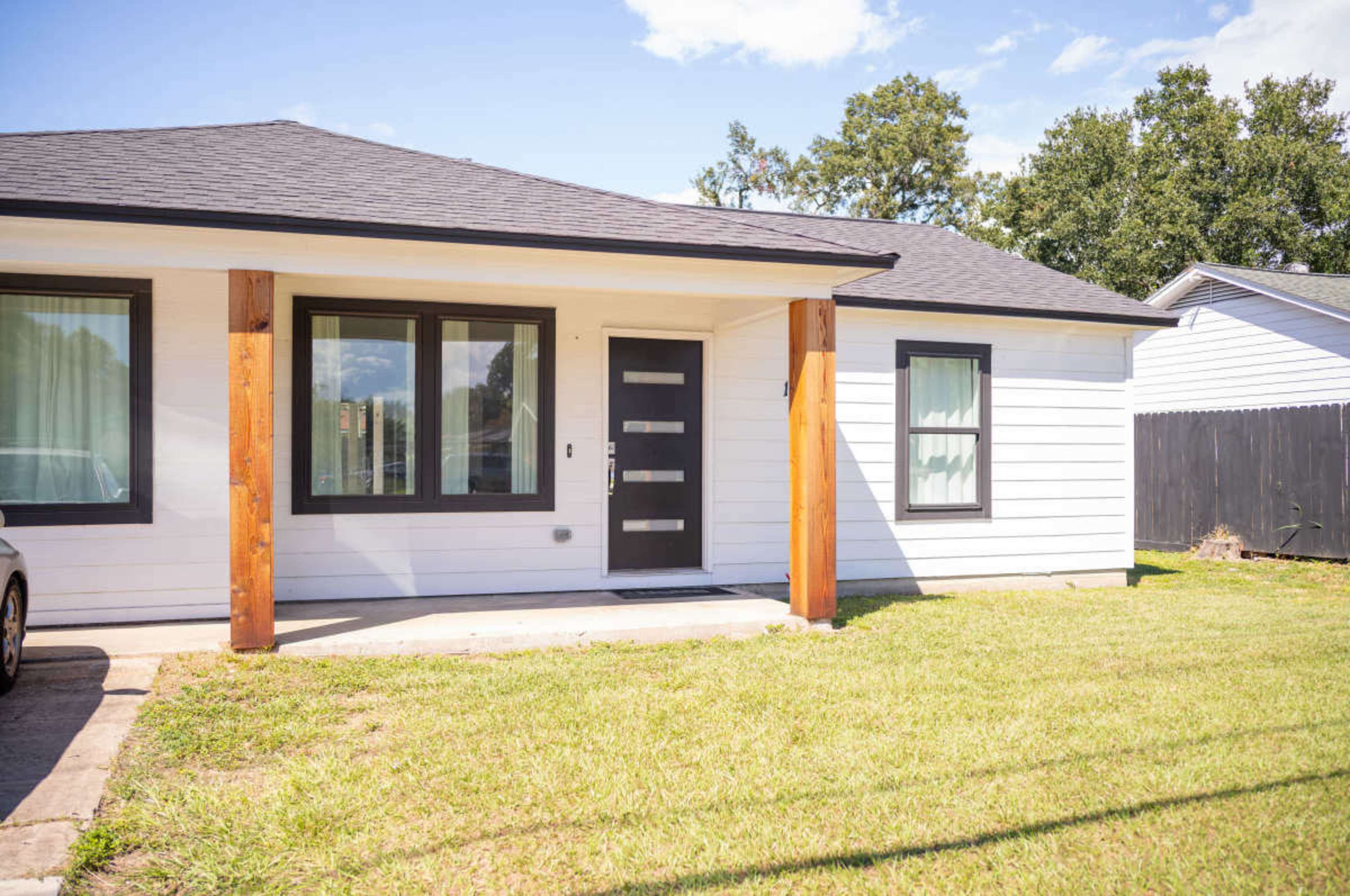 The image shows a single-story house with wooden pillars supporting a porch, large windows, and a modern front door, set in a grassy yard.