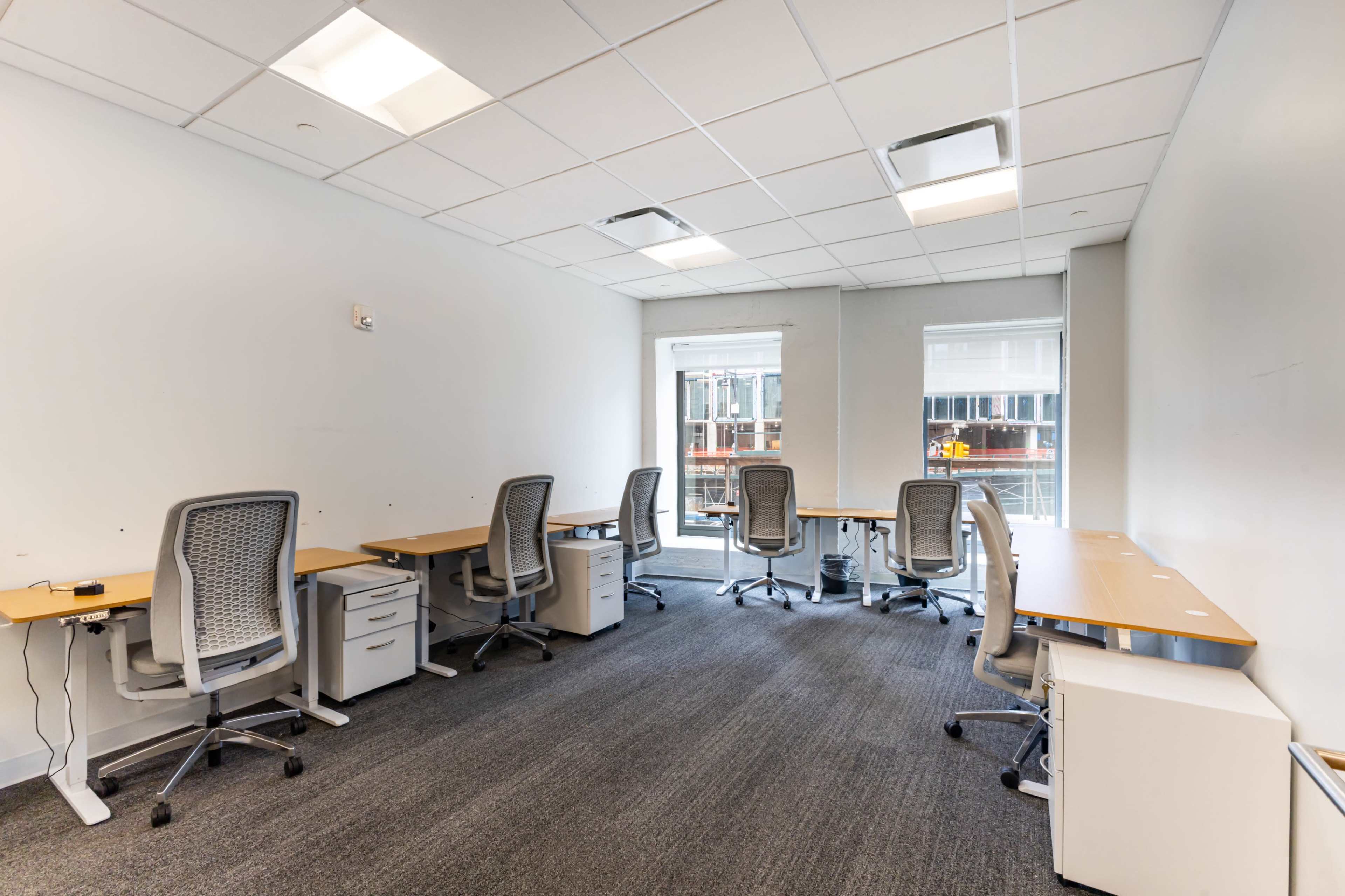 The image shows a small, empty office space with five desks and ergonomic chairs, illuminated by natural light from large windows.