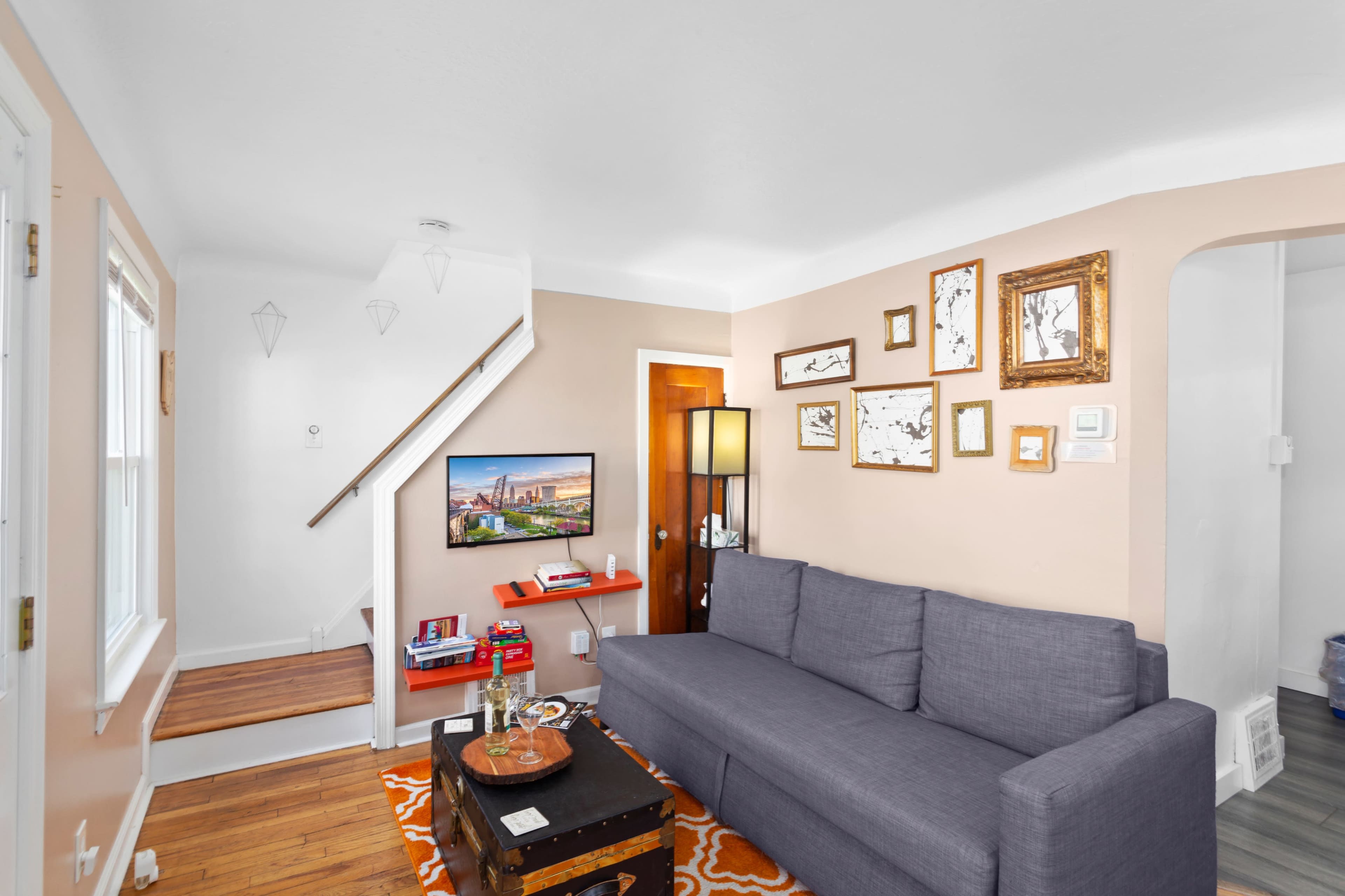 A cozy living room features a gray sofa, a coffee table, and a television mounted on the wall, with framed artwork and a staircase in the background.