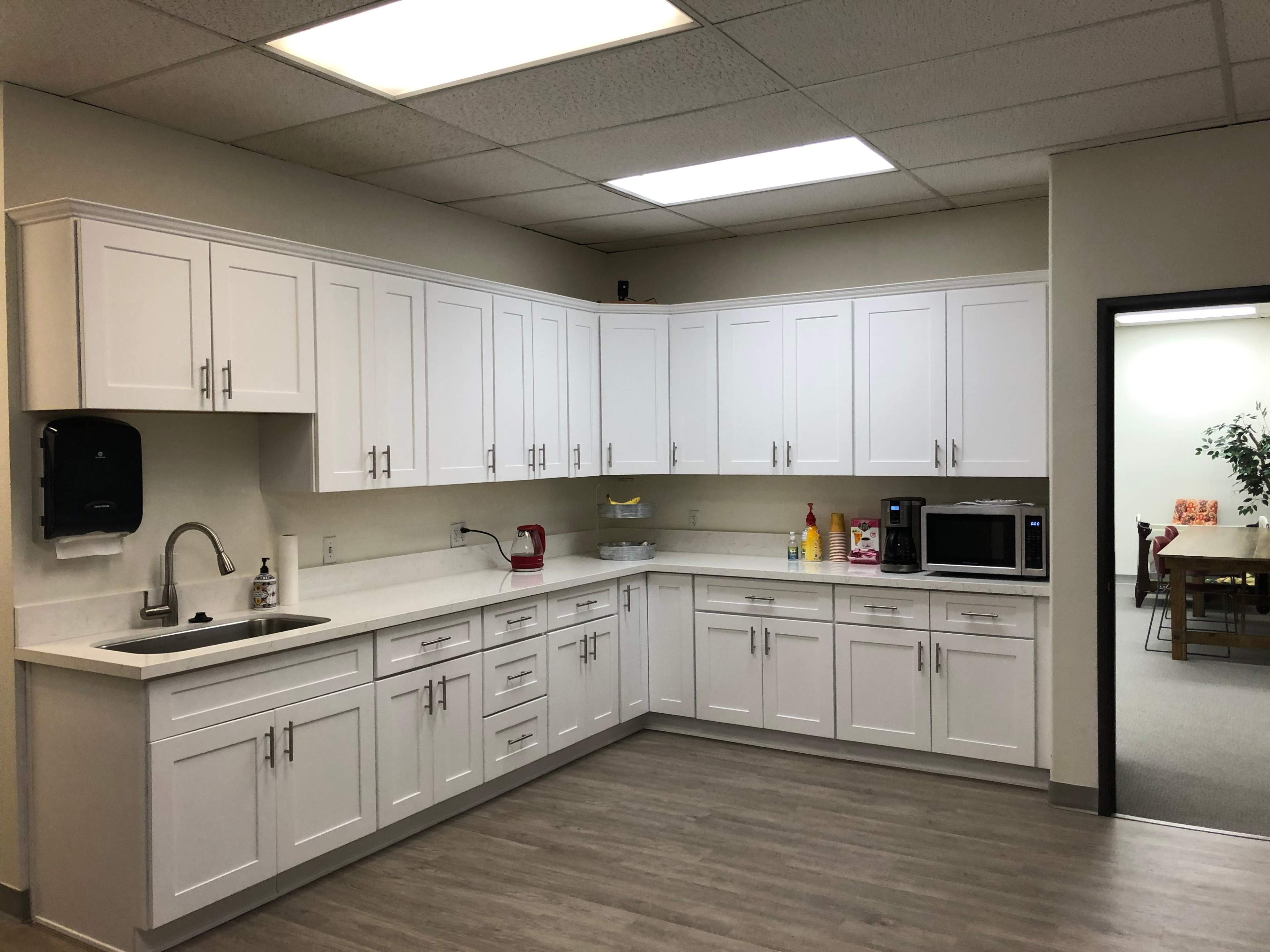 A modern kitchen with white cabinets, a sink, a microwave, and a countertop, featuring a light-colored floor and a doorway leading to another room.