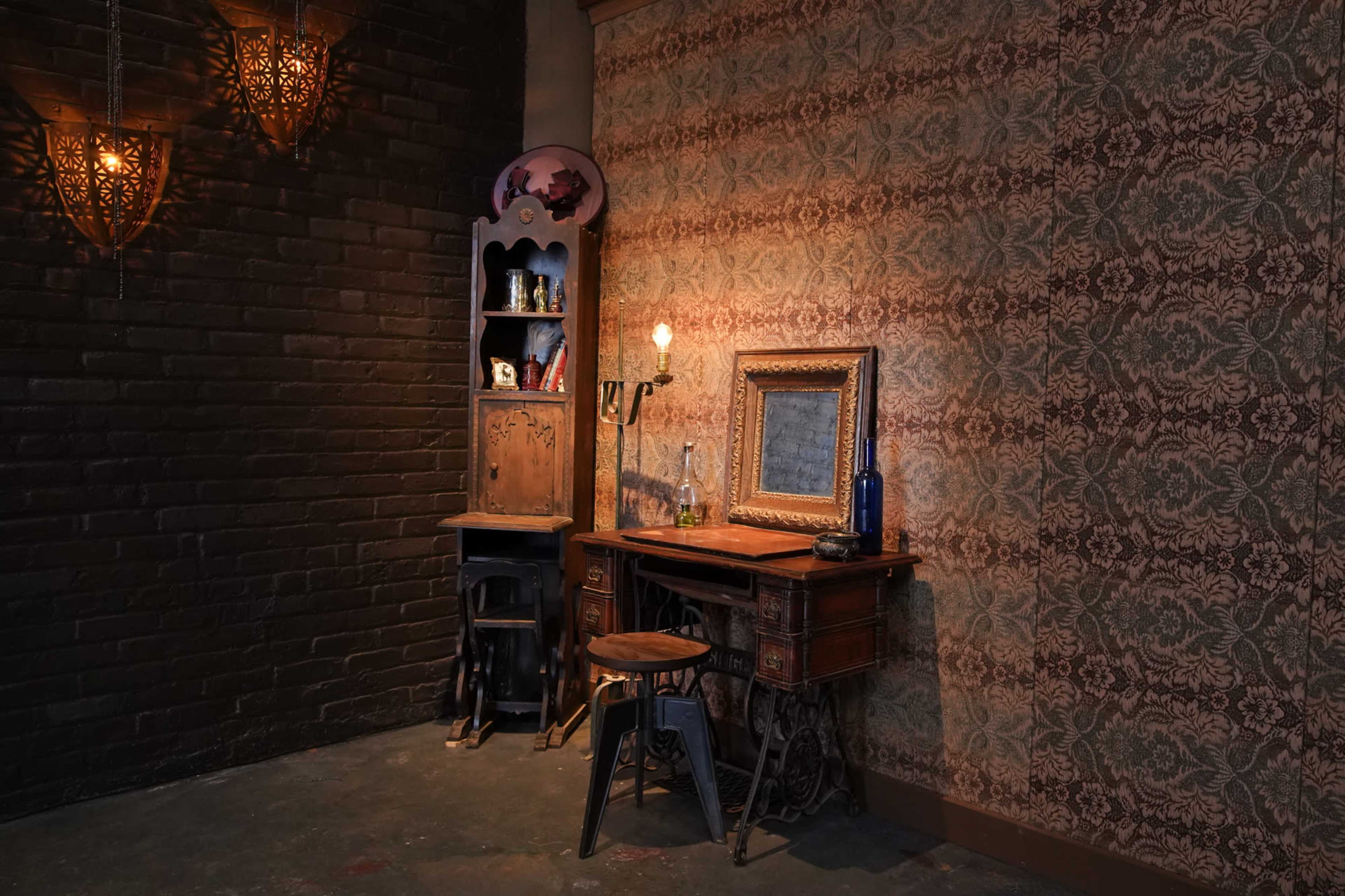 The image shows a vintage-style corner with a wooden desk, a framed mirror, a stool, and a small bookshelf against a patterned wallpaper backdrop.