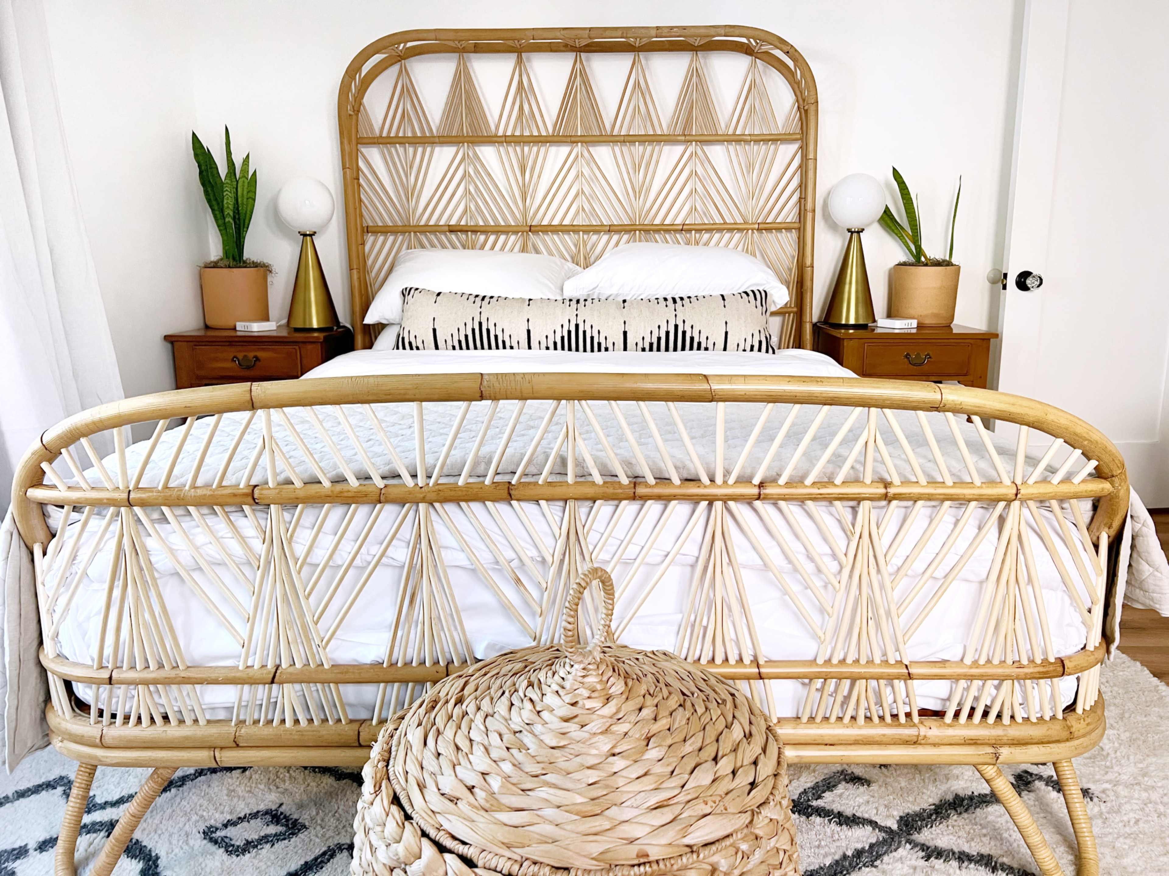 A bedroom featuring a rattan bed frame with a geometric headboard, two bedside tables, and decorative plants beside round lamps.
