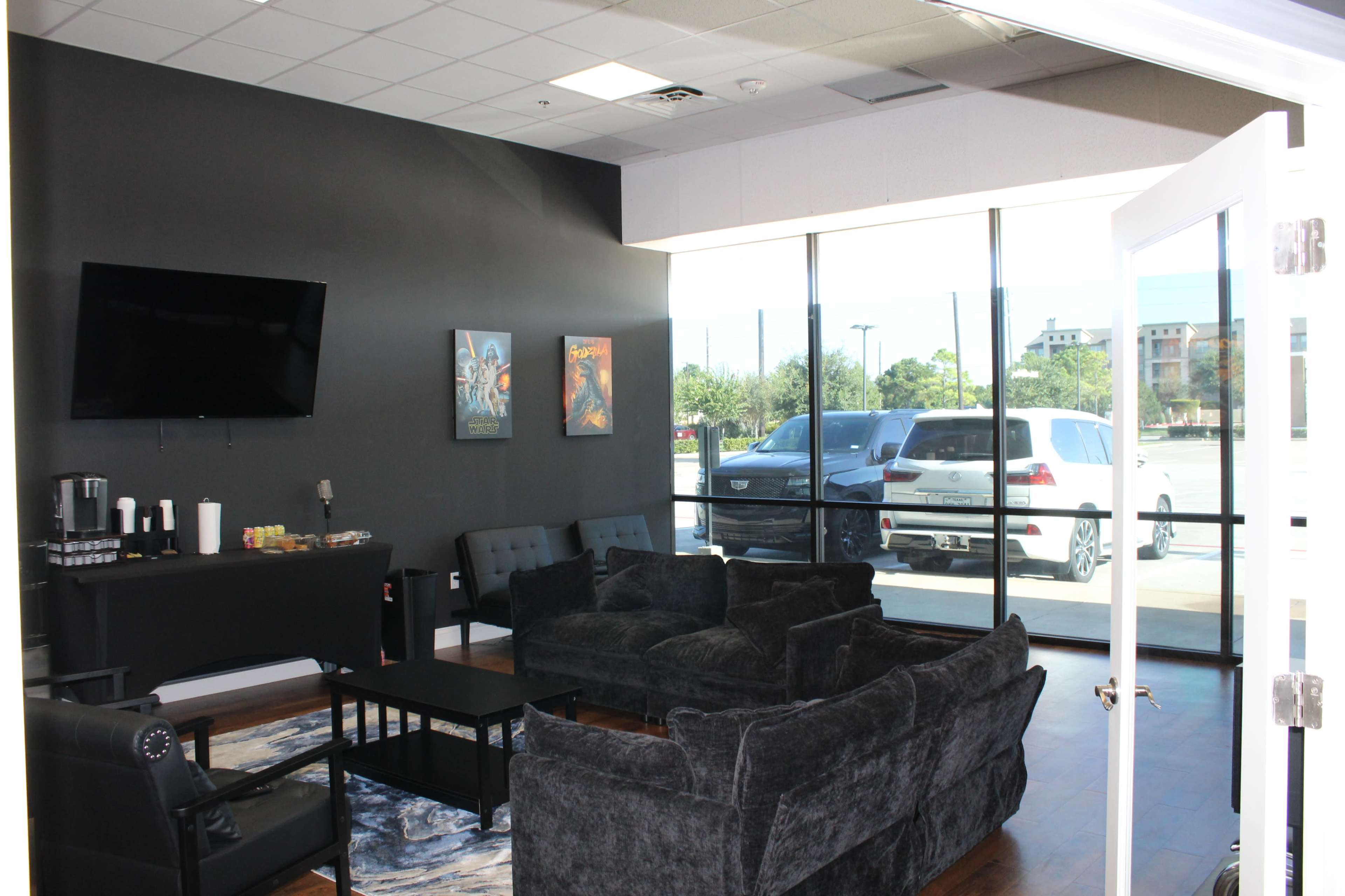 The image shows a modern waiting area with dark furniture, a television mounted on the wall, and large windows overlooking a parked car outside.
